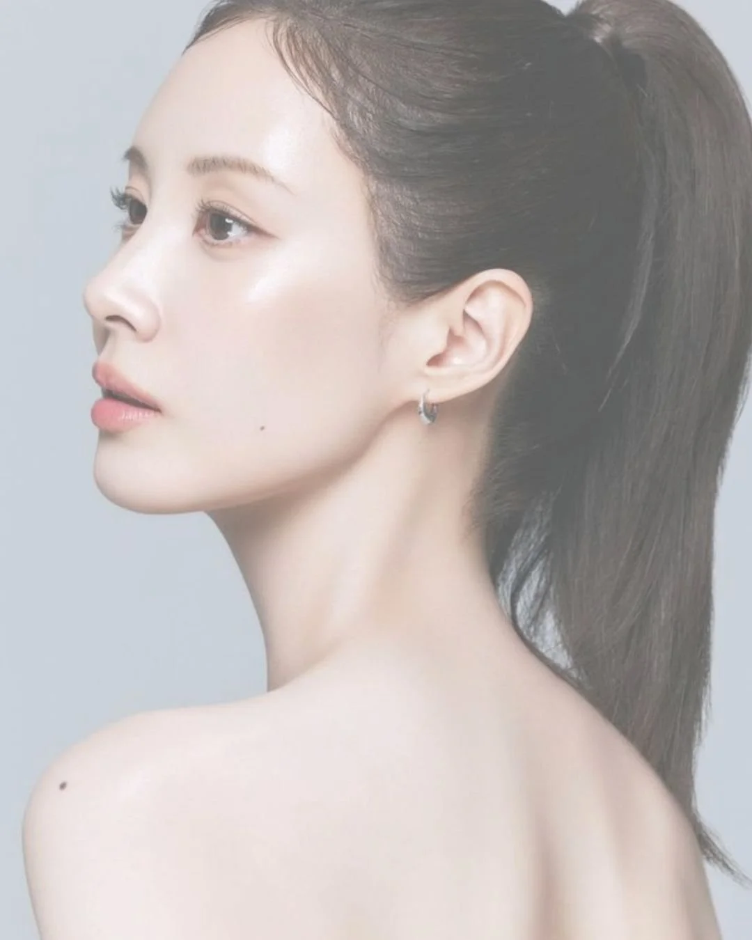 Side-profile portrait of a woman with a dark ponytail and hoop earring against a light gray background