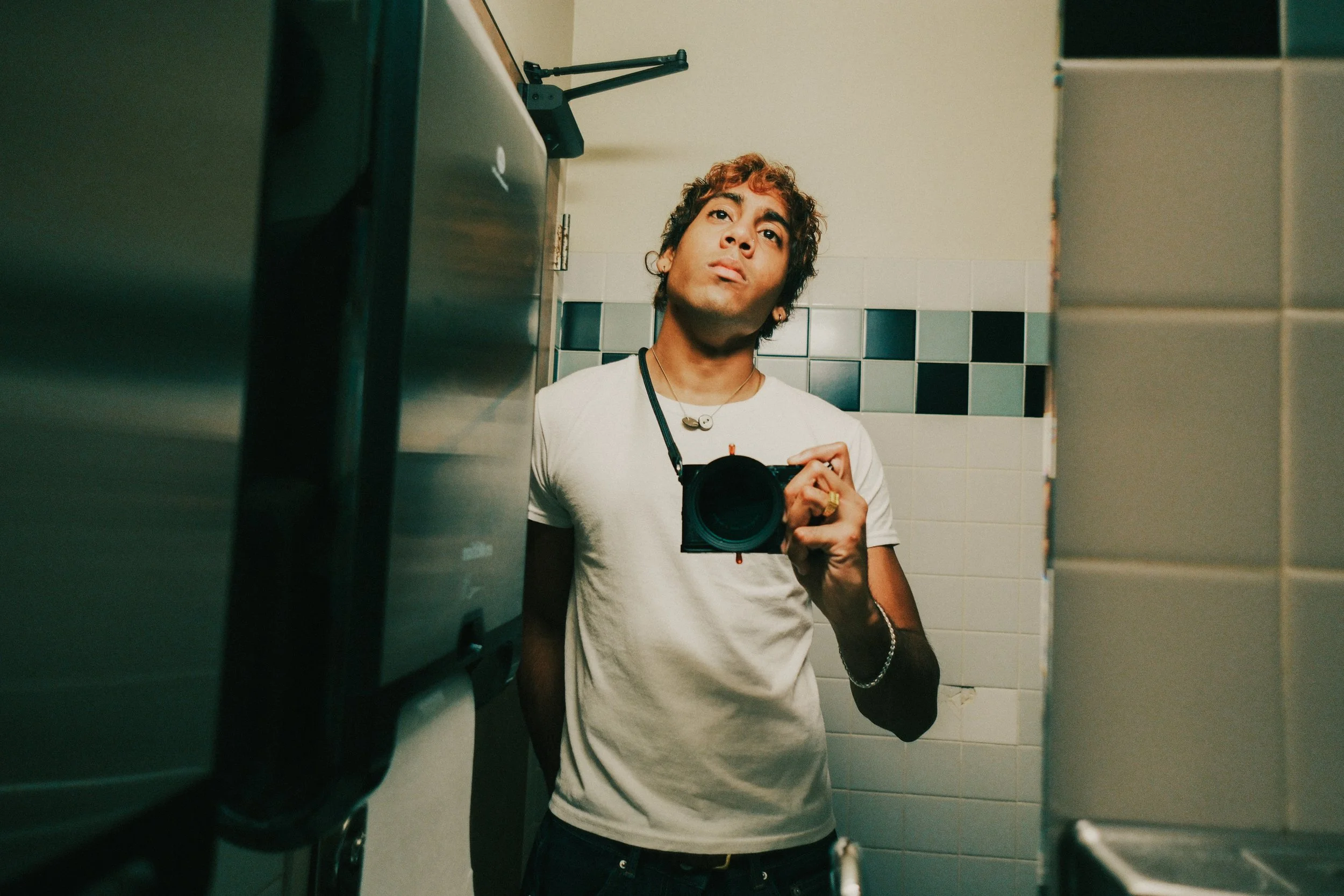 A young man with curly hair and earrings taking a mirror selfie in a bathroom, holding a camera, wearing a white t-shirt and a necklace.