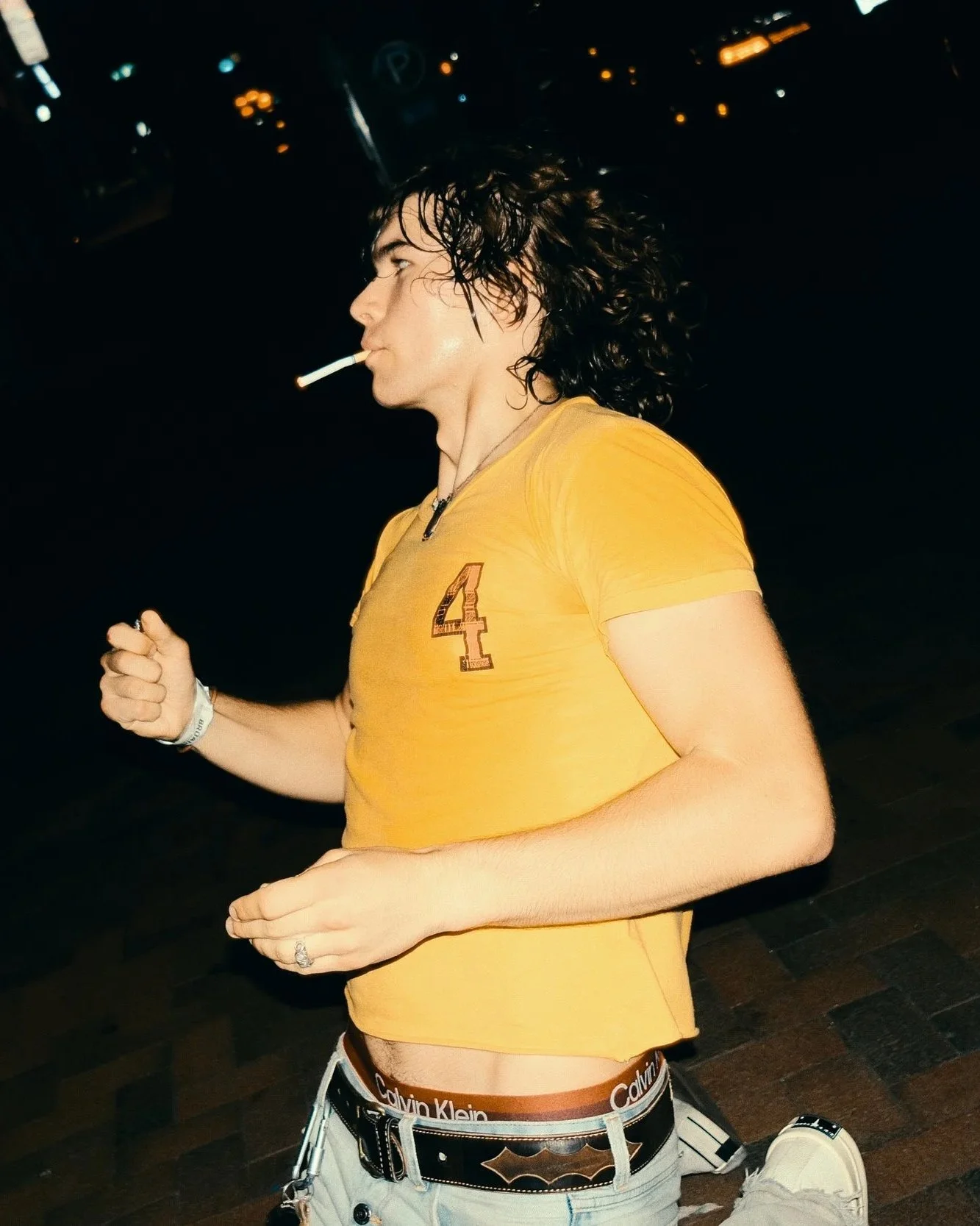 A person with long, wet, curly hair running in a dark setting, wearing a yellow t-shirt with the number 4 on it, light-colored shorts, and a black Batman belt.