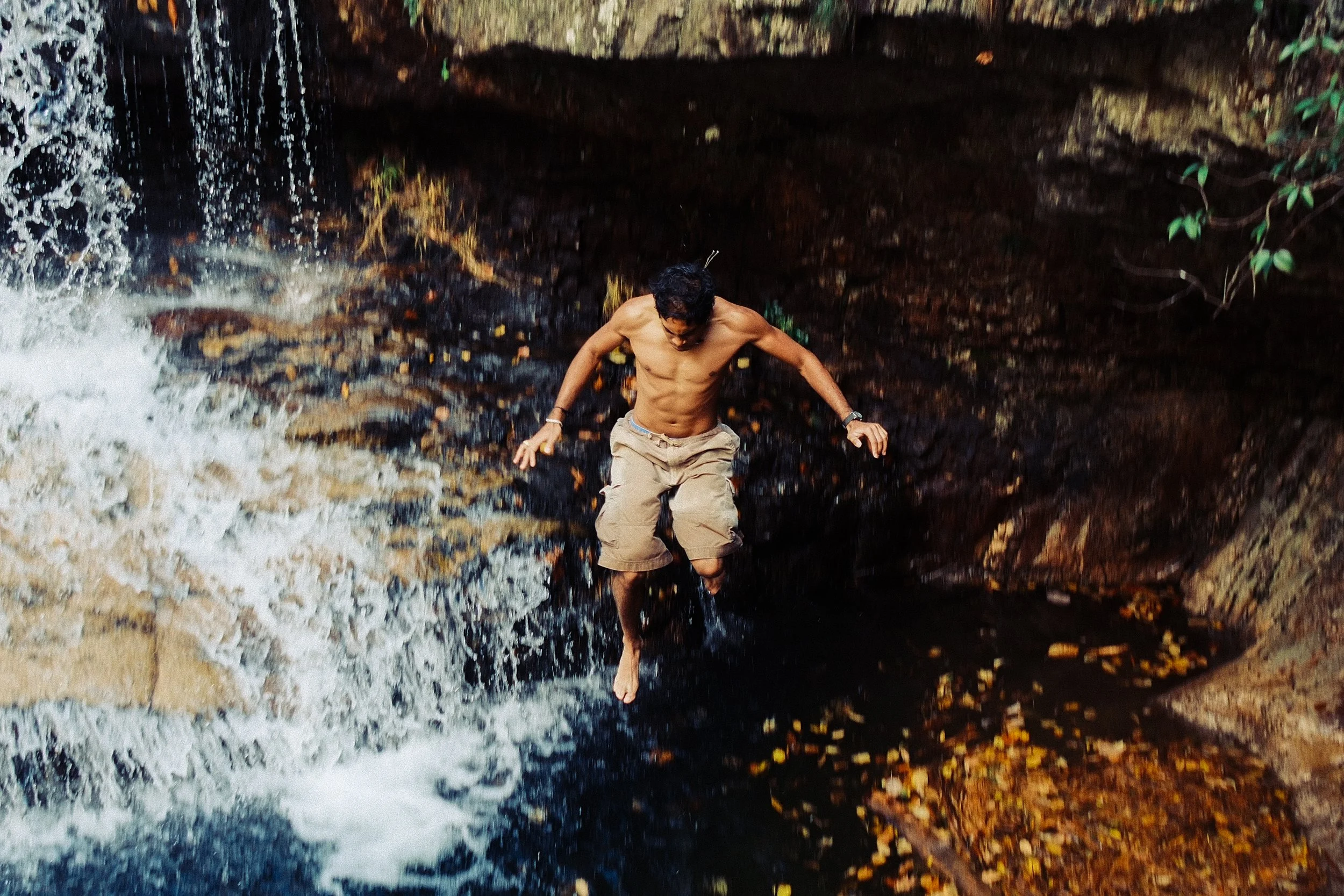 A shirtless man with dark hair standing under a small waterfall or stream in a rocky area surrounded by dark rock and greenery.