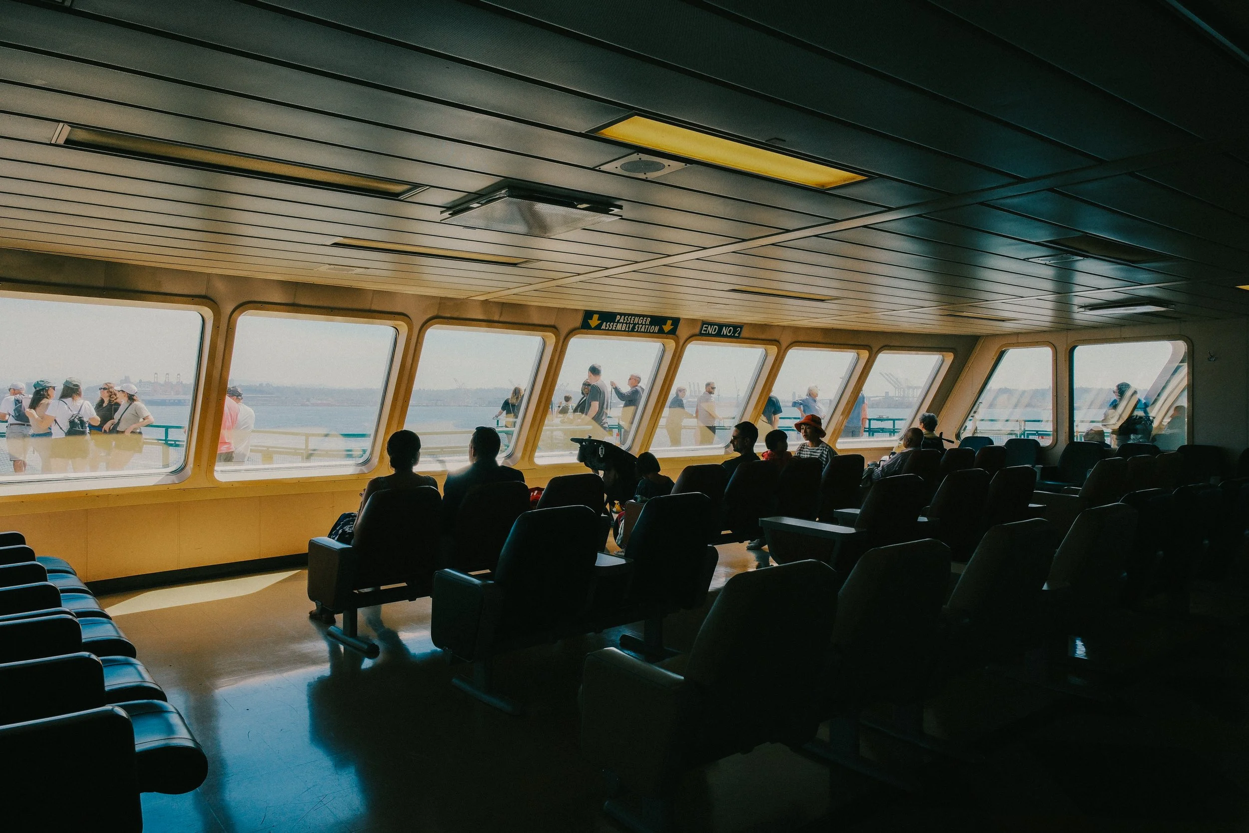 Indoor view of a ferry terminal waiting area with black chairs, large windows showing people outside near the water, and a sign indicating passenger assembly station and end number 2.