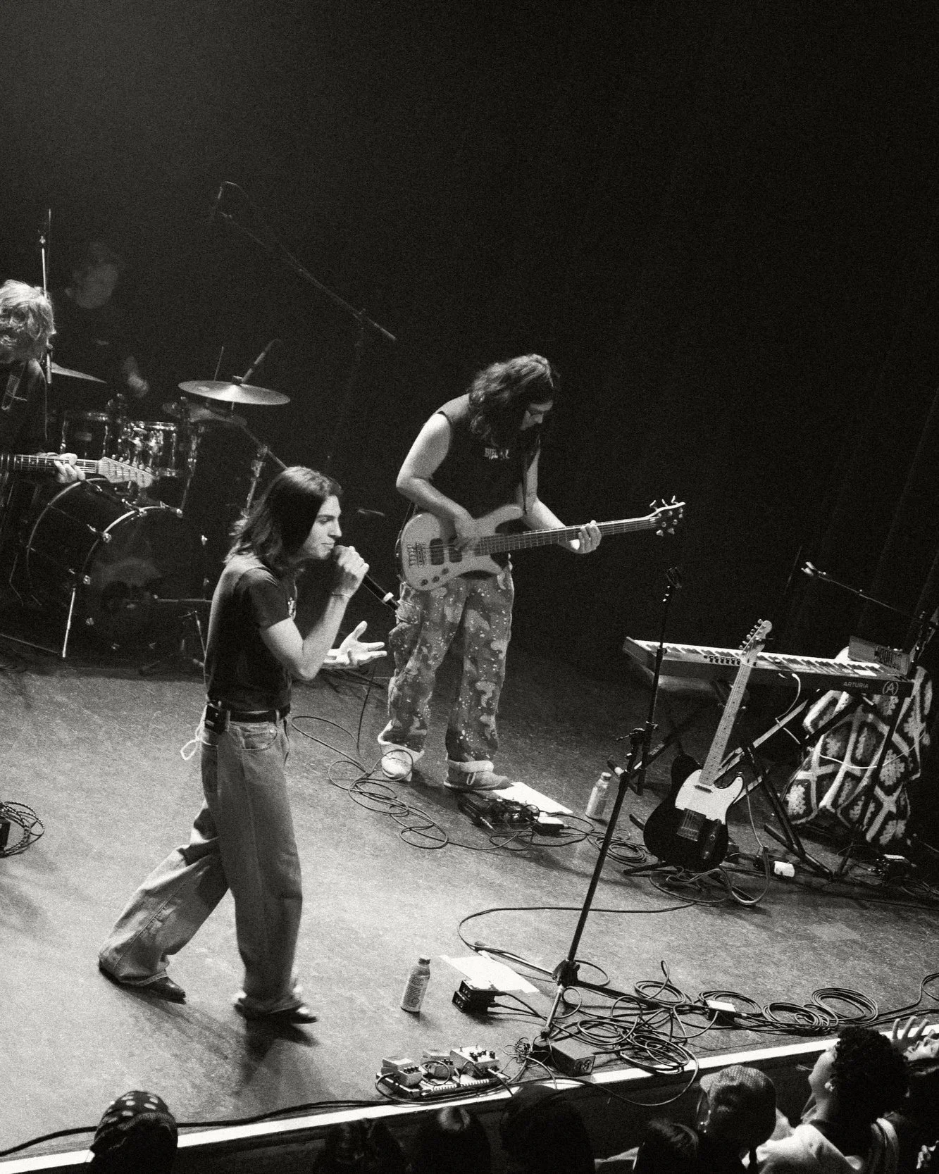 Black and white photo of a live band performance on stage, featuring a female singer with long hair in a dark t-shirt and jeans, a guitarist with long hair in camouflage pants, and a drummer in the background. Various musical equipment and cables are visible on stage.