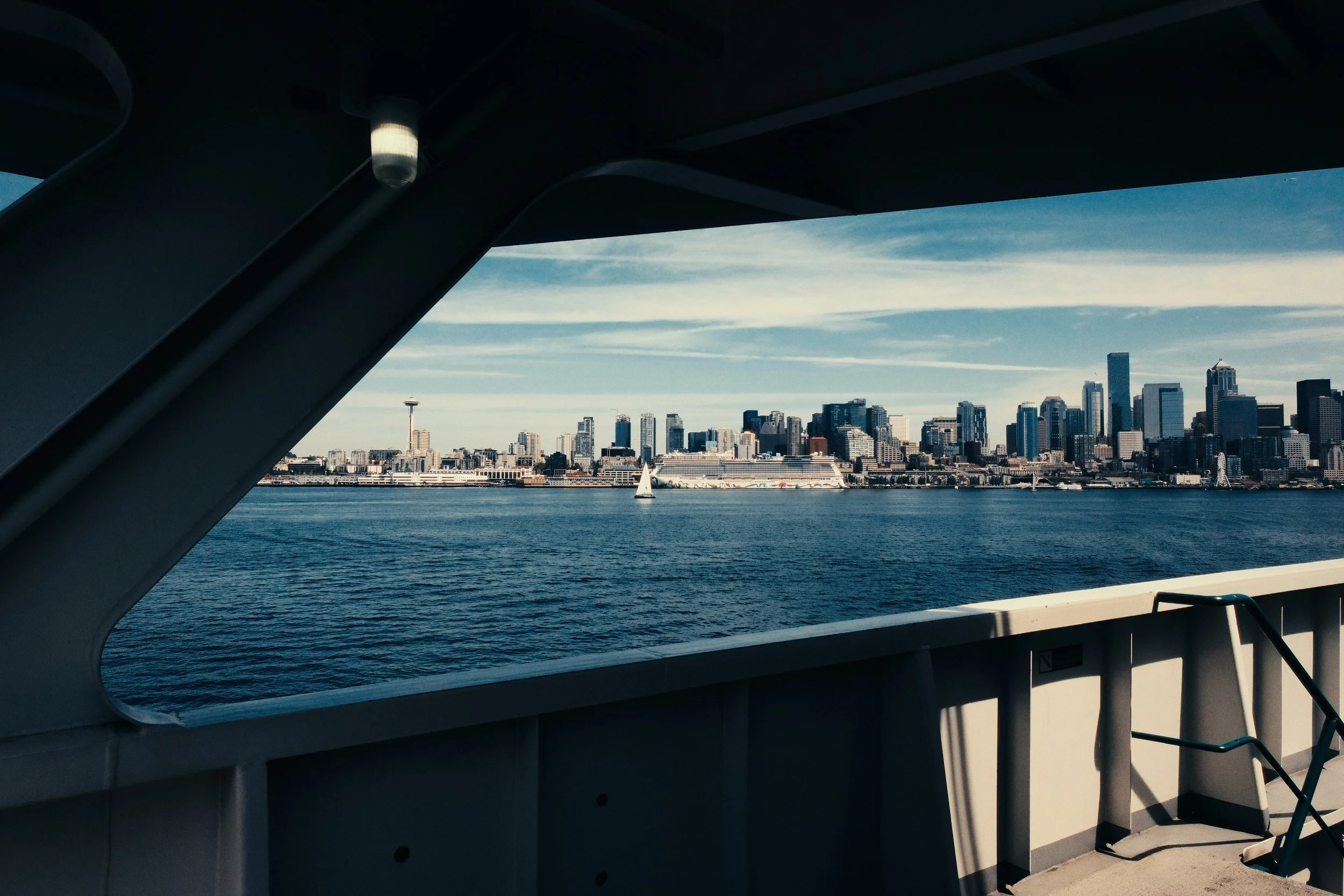 View of Seattle skyline from a boat, with the Space Needle visible, across the water with a sailboat in the distance, framed by a highway bridge.