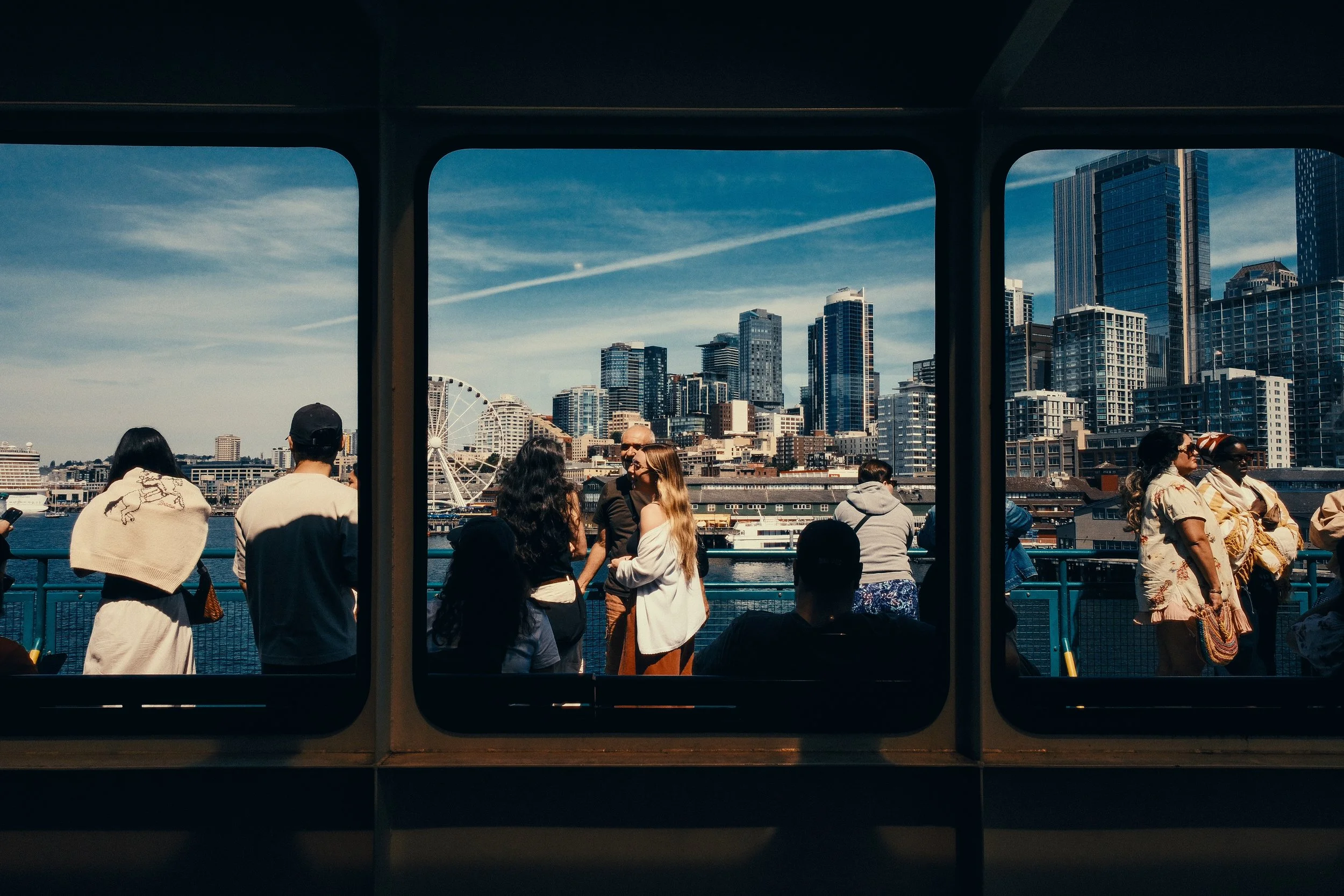 People standing and sitting on a ferry overlooking a city skyline with tall buildings and a ferris wheel, framed by the ferry window.