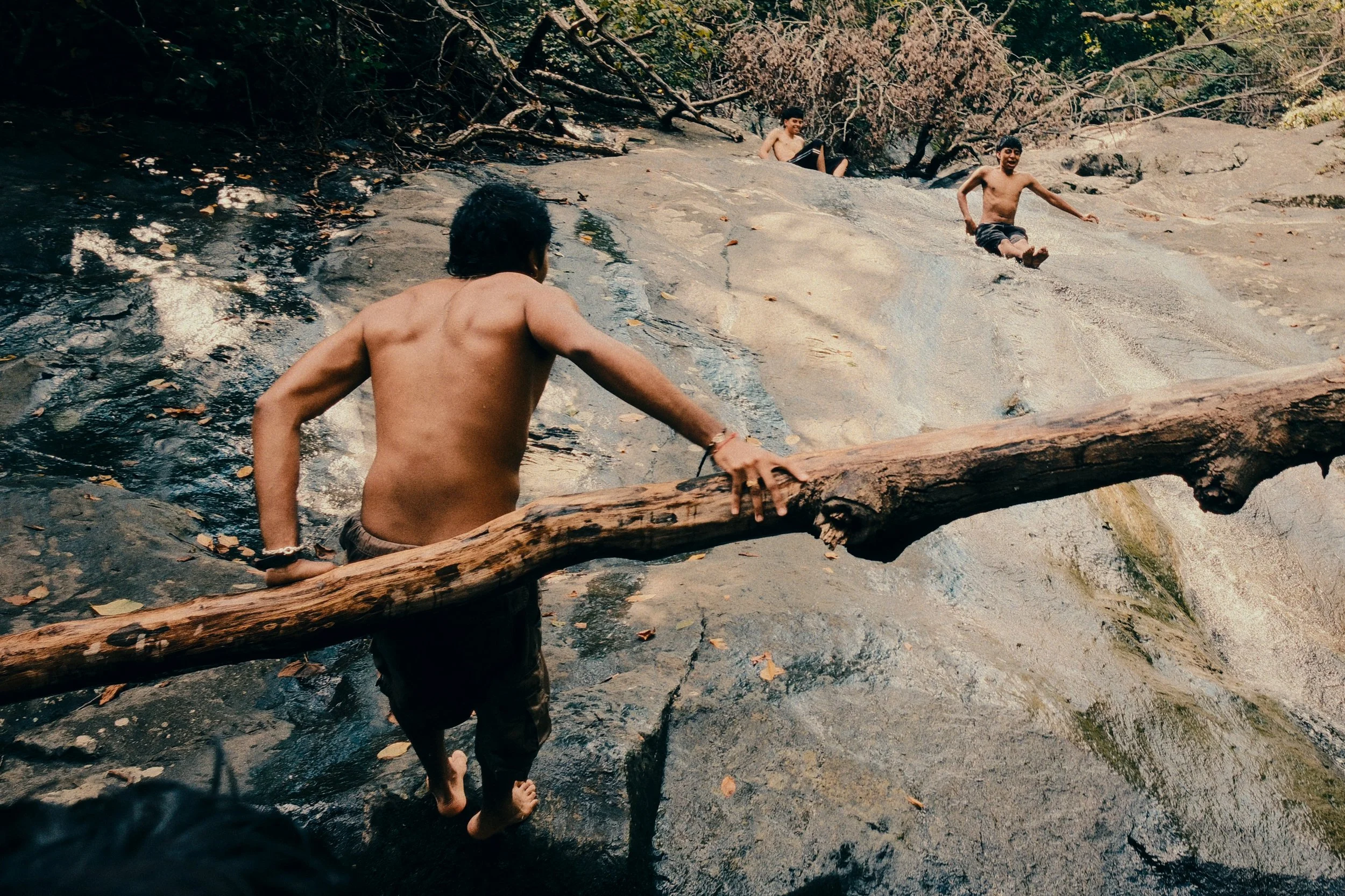 Three boys playing near a rocky stream in a wooded area, one of them climbing a large rock, two others walking through the water.