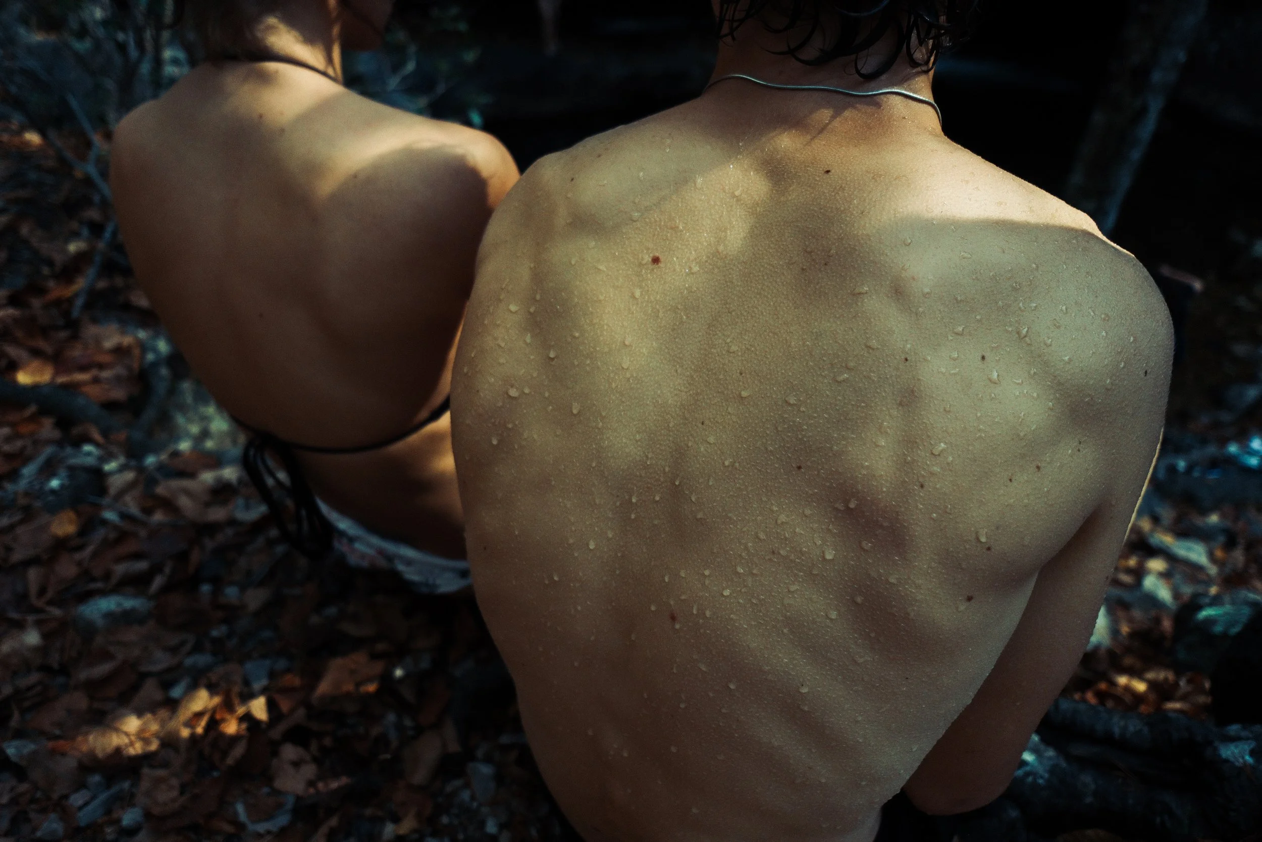 Close-up of two bare human backs with wet skin and water droplets, outdoors in a wooded area.