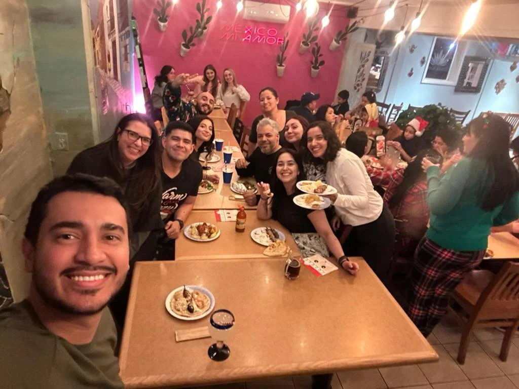 Large group posing for a selfie at a lively Mexican restaurant surrounded by tables, plates of food, and pink decor.