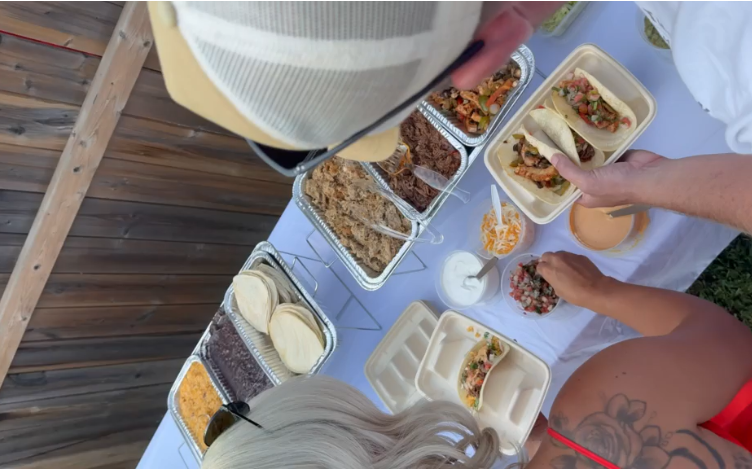 Overhead view of a taco buffet with tortillas, seasoned meats, rice, beans, salsa, cheese, and toppings being served outdoors.