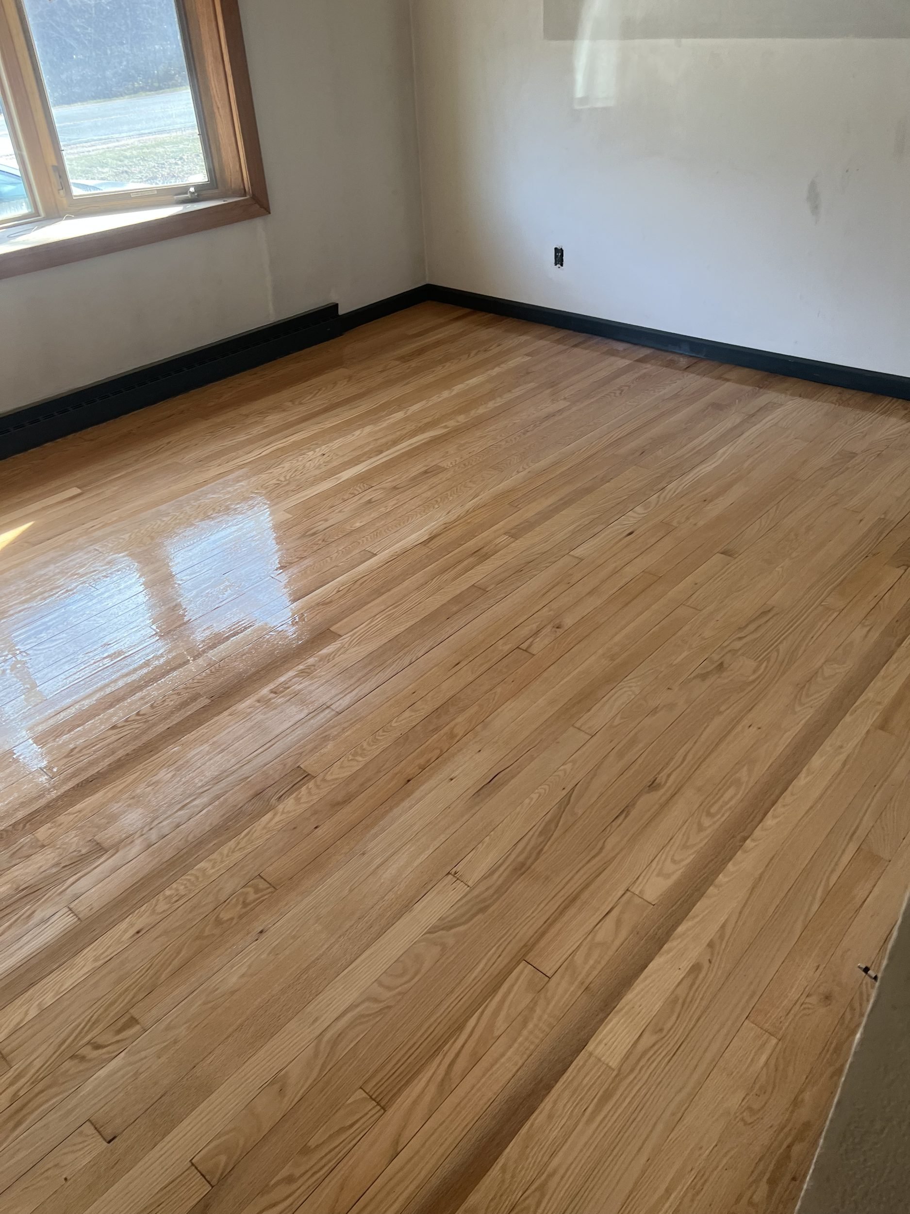 Empty room with hardwood floor, white walls, a window with wooden trim, and black baseboard heating.