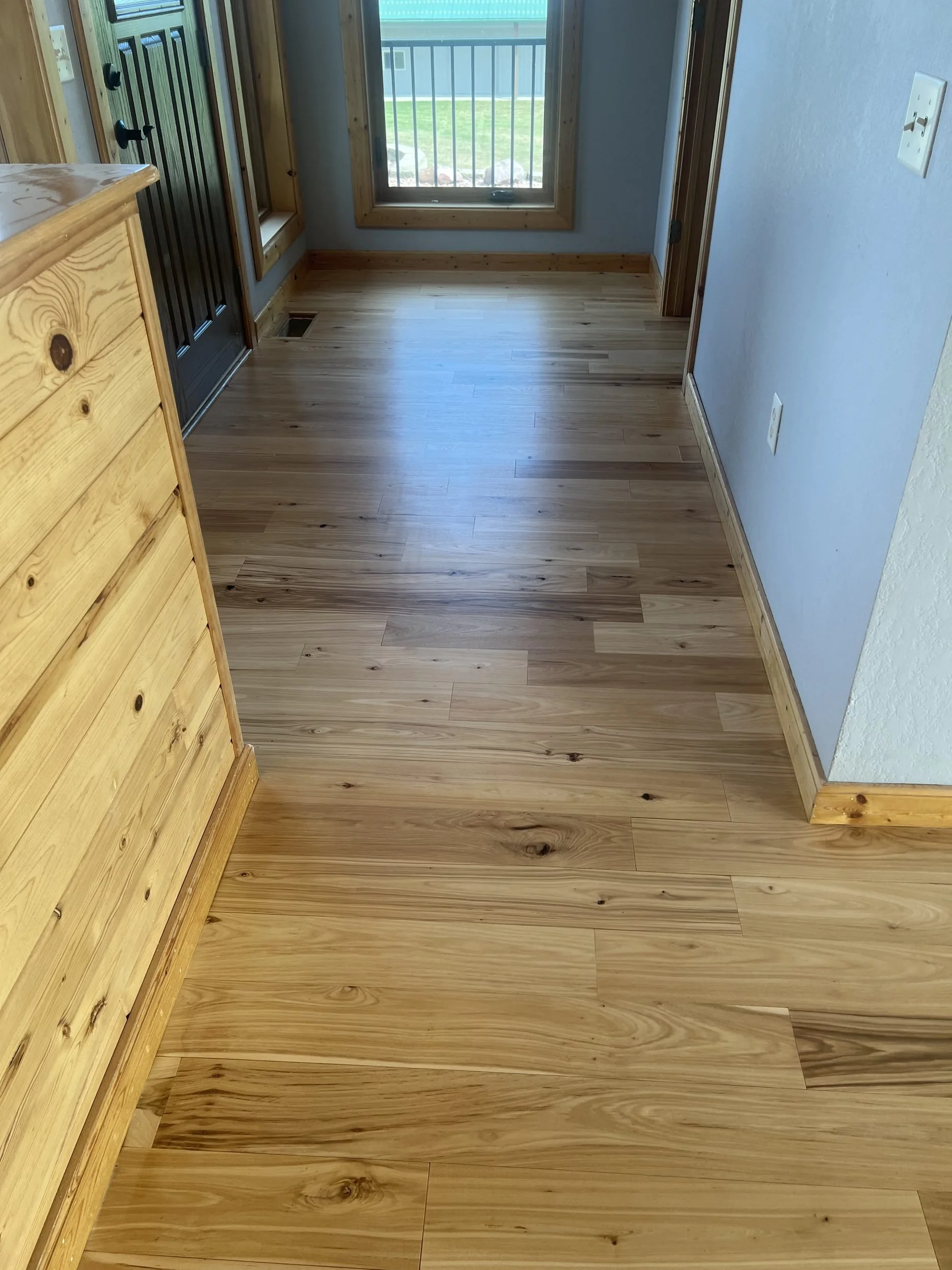 A hardwood floor in a room with light-colored wood flooring and a sliding glass door leading to a small balcony or patio with a view of a grassy yard and a fence.