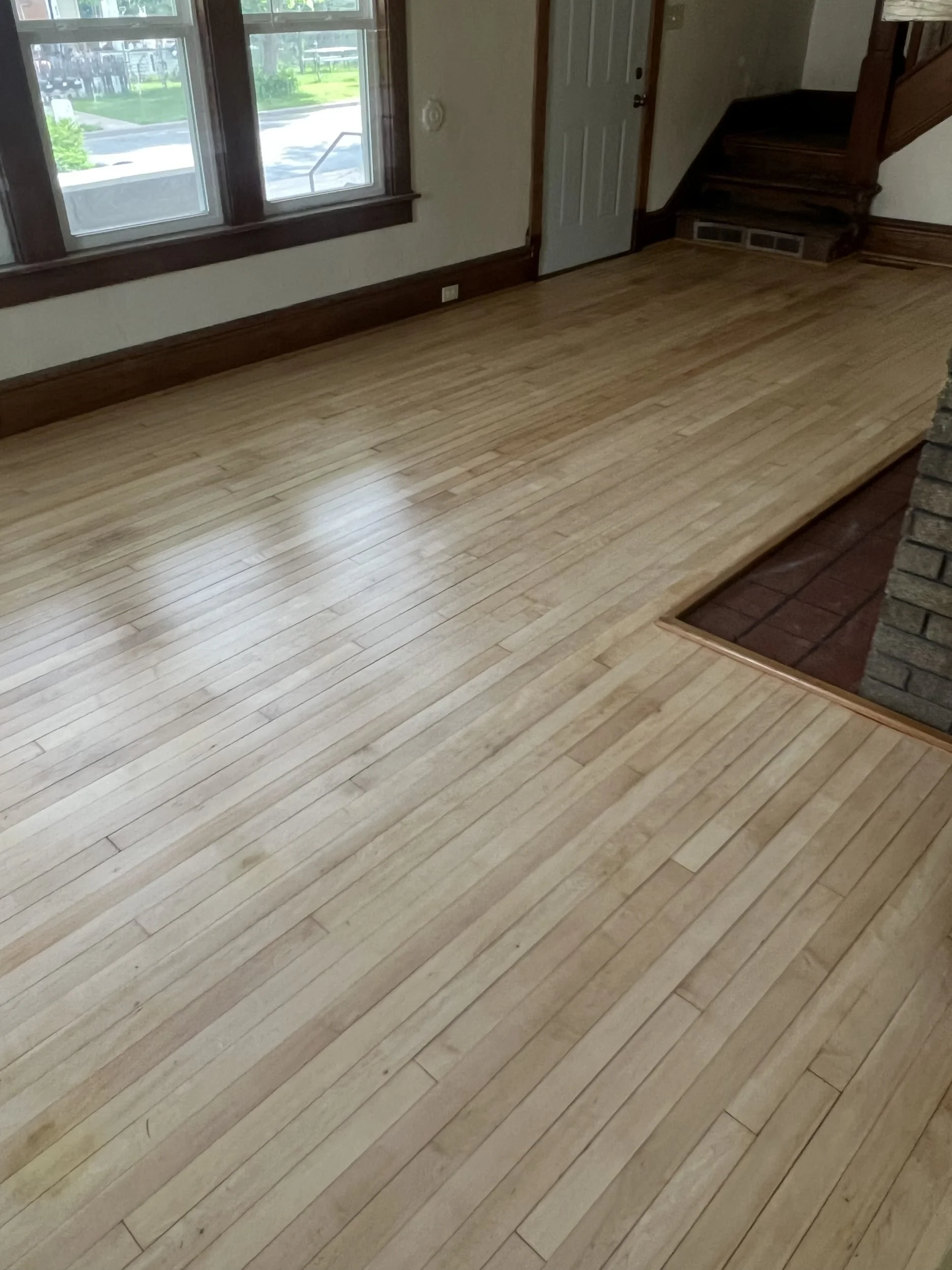 Empty room with hardwood floor, large window, wooden trim, door, and staircase with wood railing.