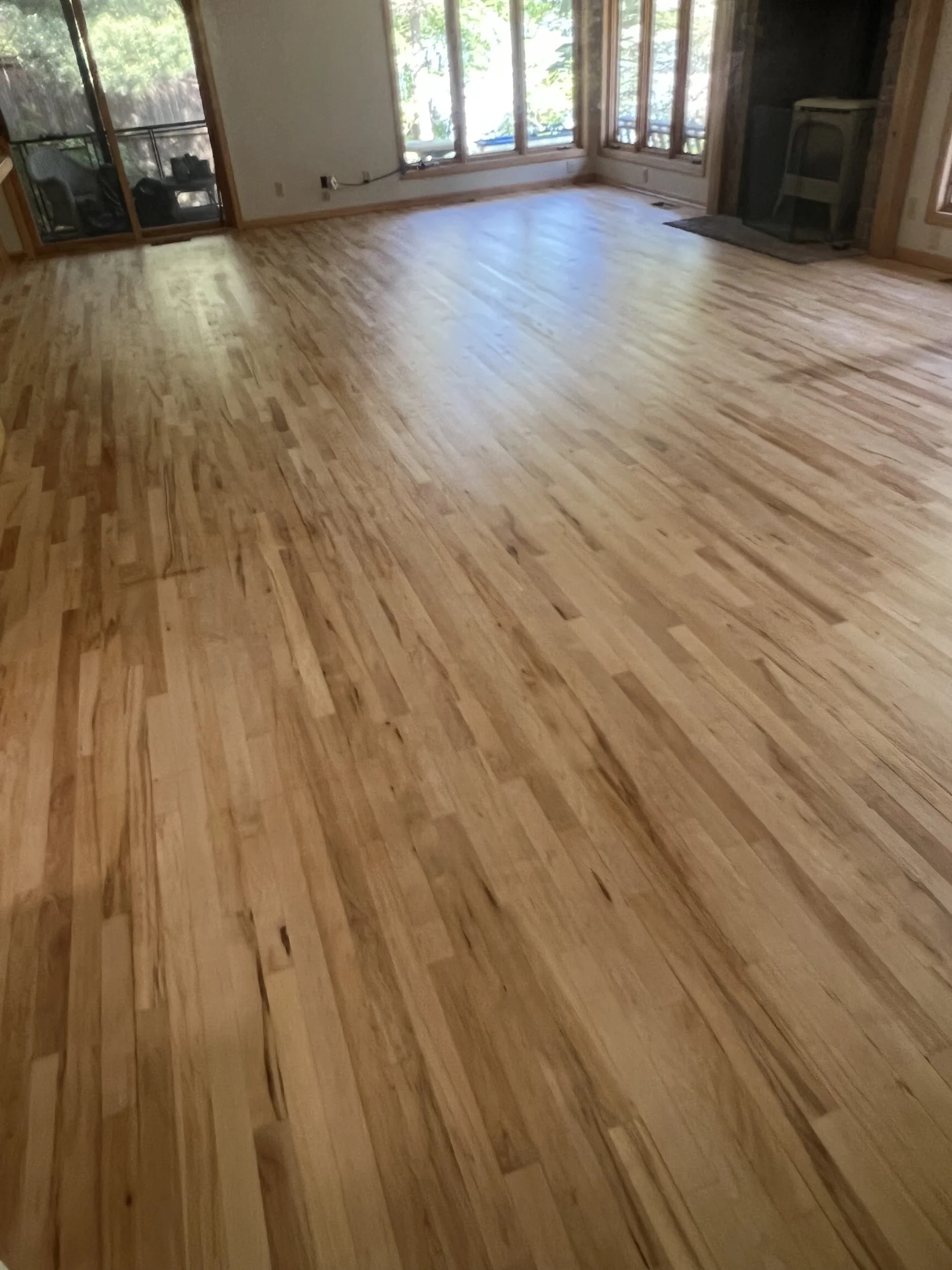 A room with newly installed hardwood flooring, large windows, a sliding glass door, and a wood-burning stove in the corner.