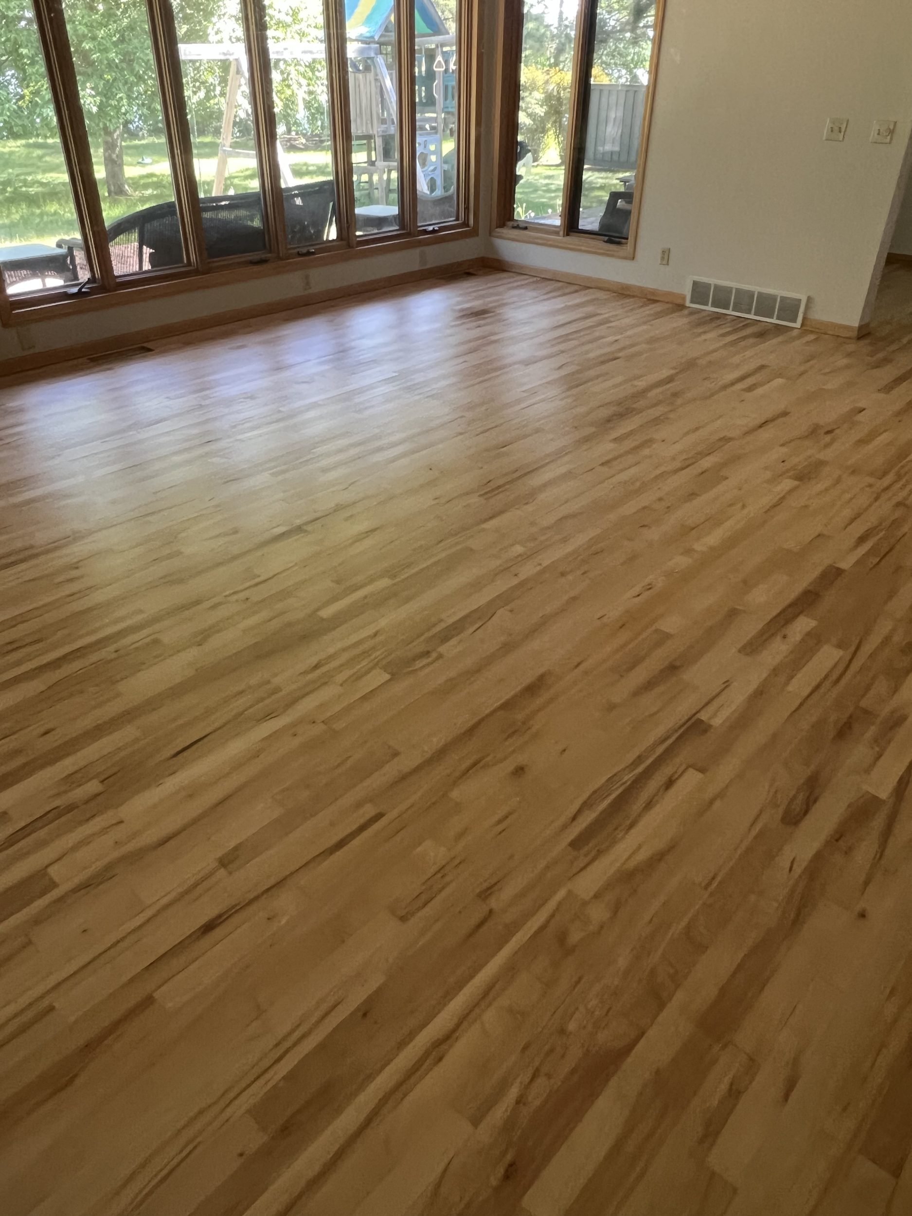 Empty living room with hardwood floors, large windows, and a view of a backyard with trees, outdoor furniture, and a playground set.