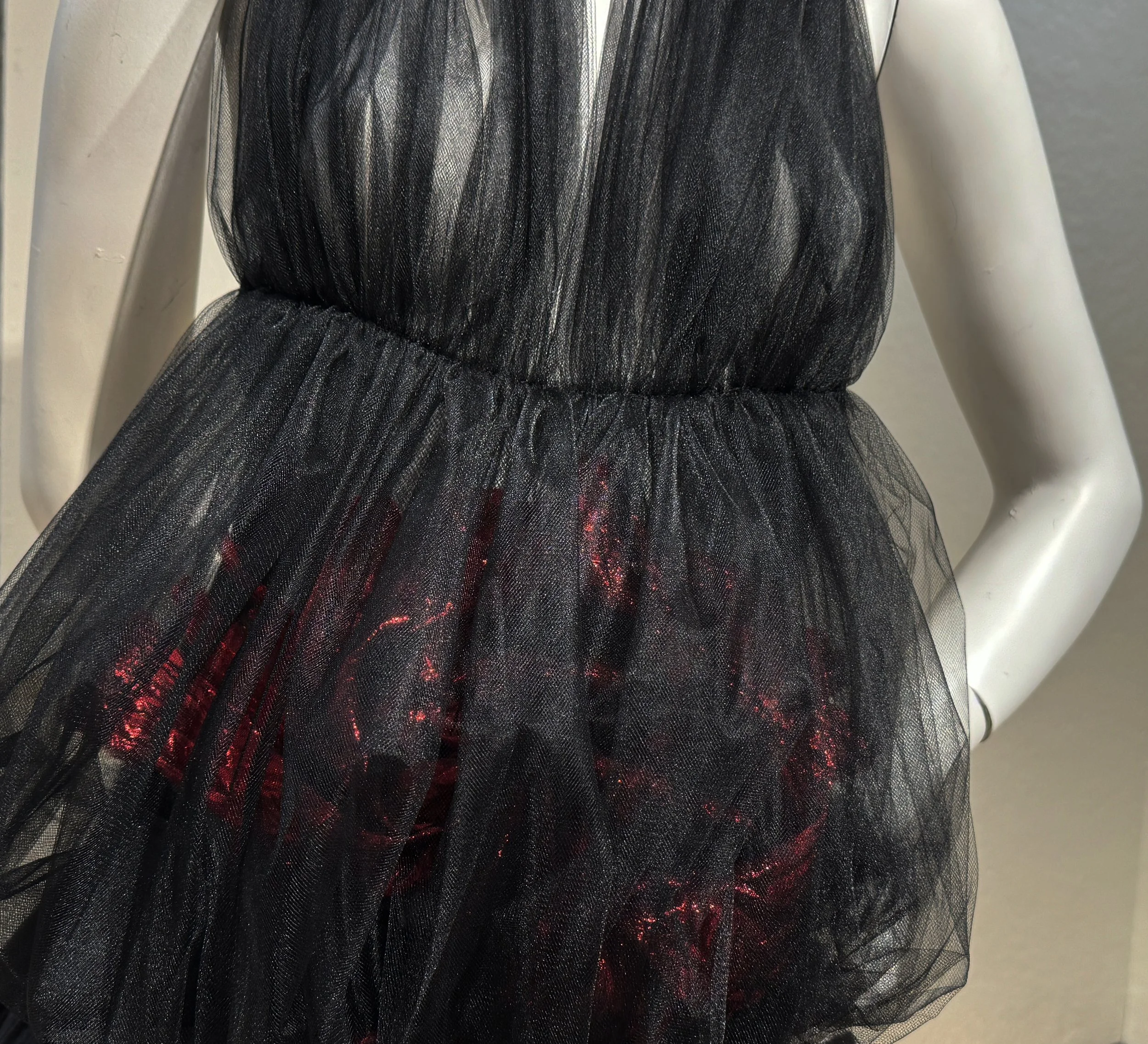 Close-up of a black sheer tulle dress with a gathered waist and red glittery fabric underneath on a mannequin