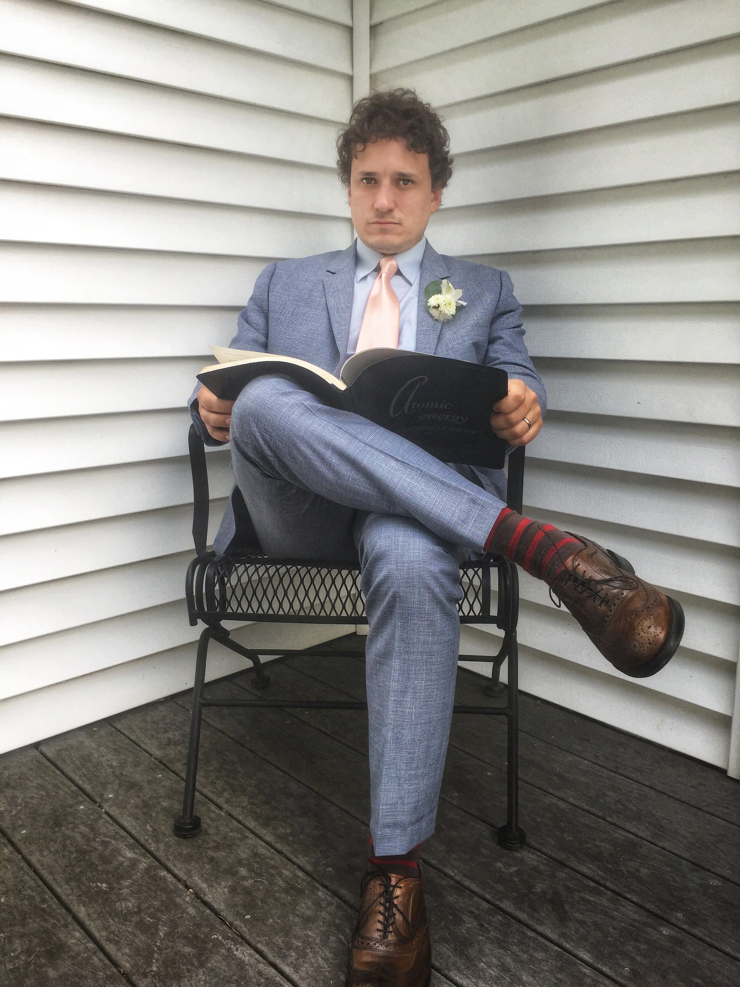 Man in a blue suit and pink tie with a boutonniere, seated cross-legged and reading a book outdoors.