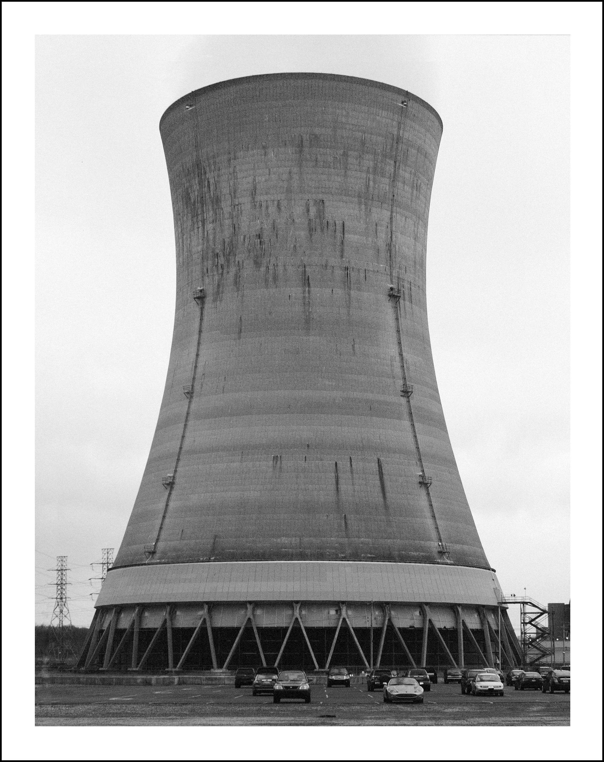 Black-and-white photo of a large concrete cooling tower with cars parked in front and power transmission towers nearby