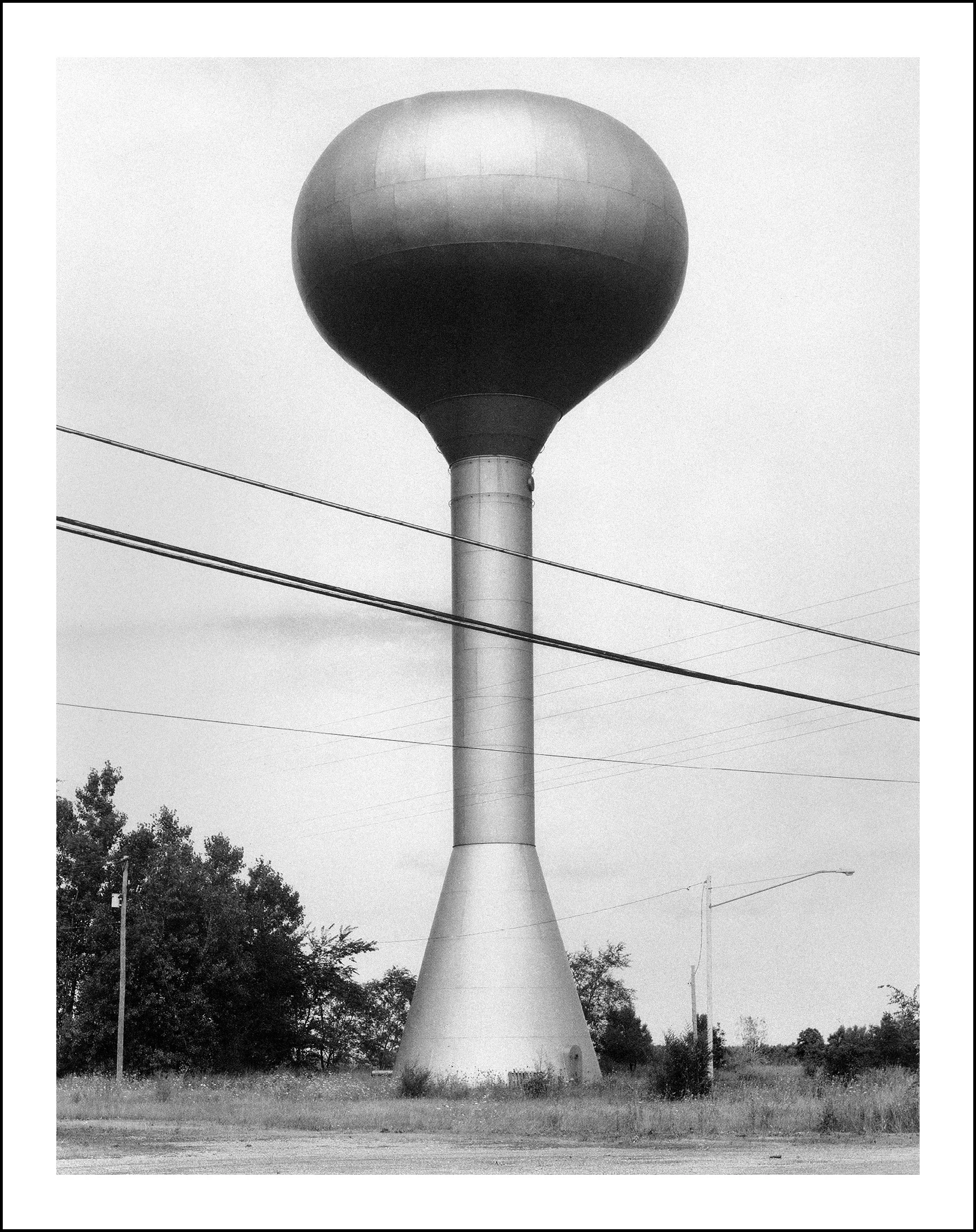 Black-and-white photo of a tall water tower with a spherical tank, utility wires, trees, and roadside grass