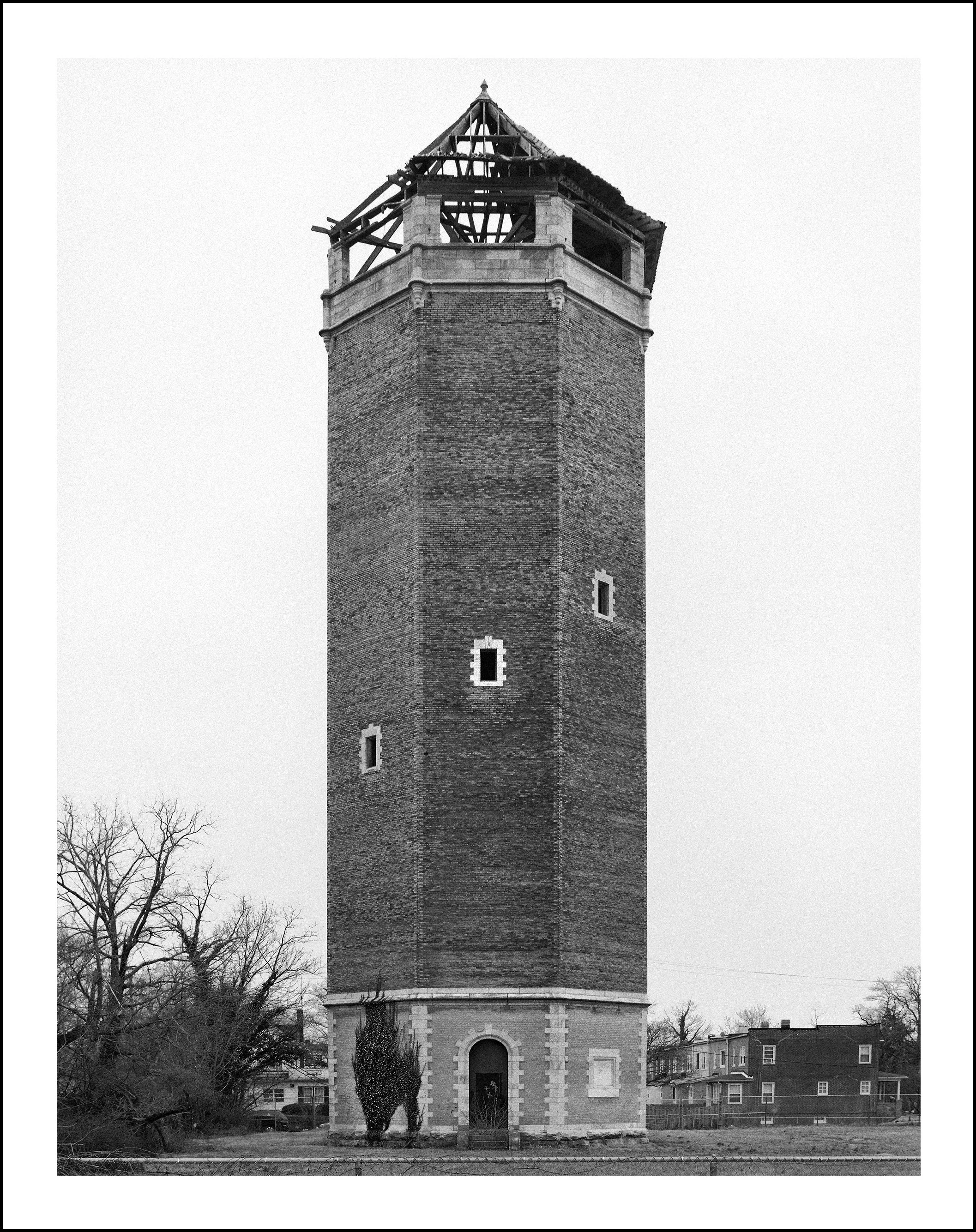 Black-and-white photograph of a tall historic brick church tower with a partially collapsed wooden roof, surrounded by leafless trees and houses.