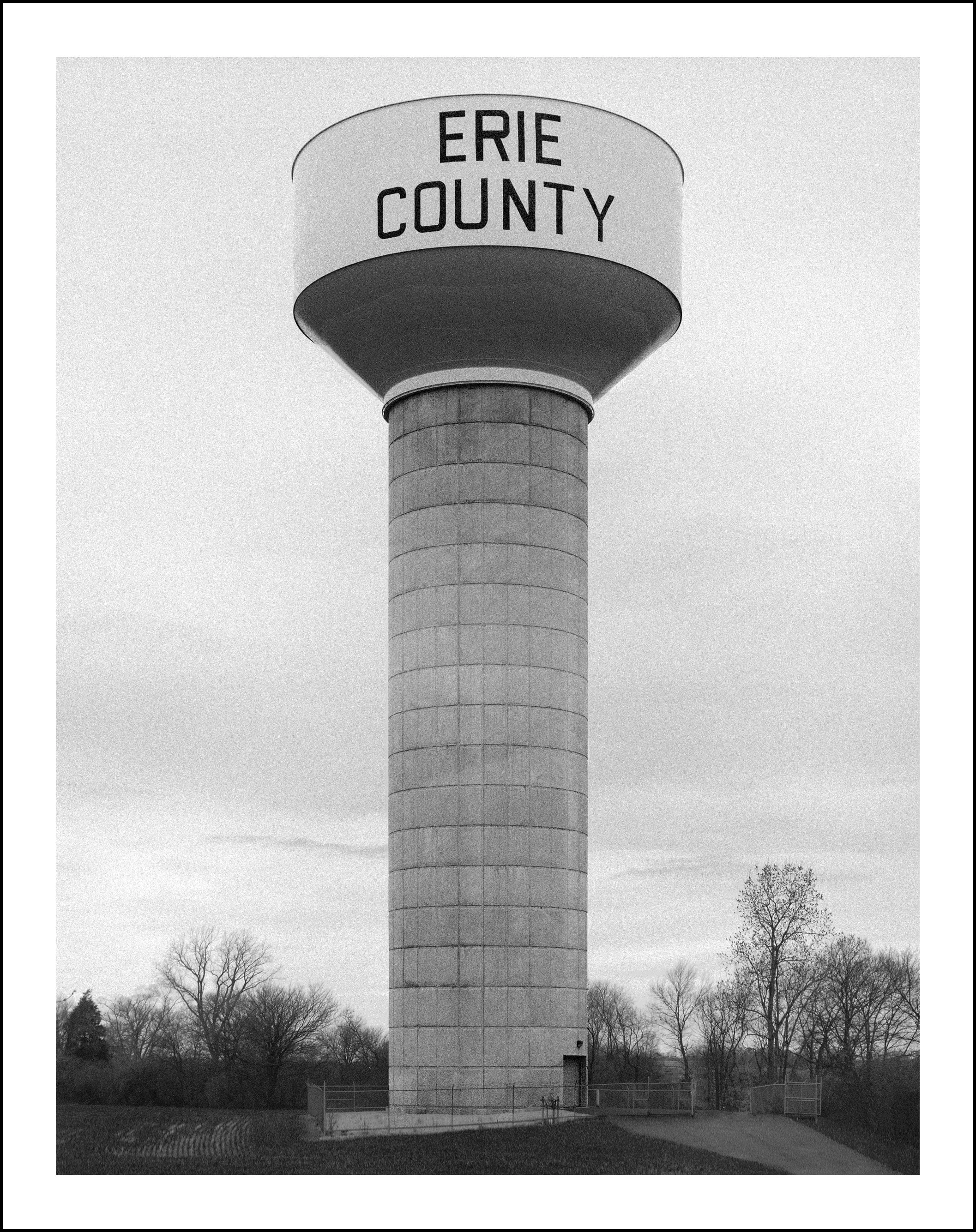 Black-and-white photo of a tall water tower labeled “Erie County” surrounded by trees.