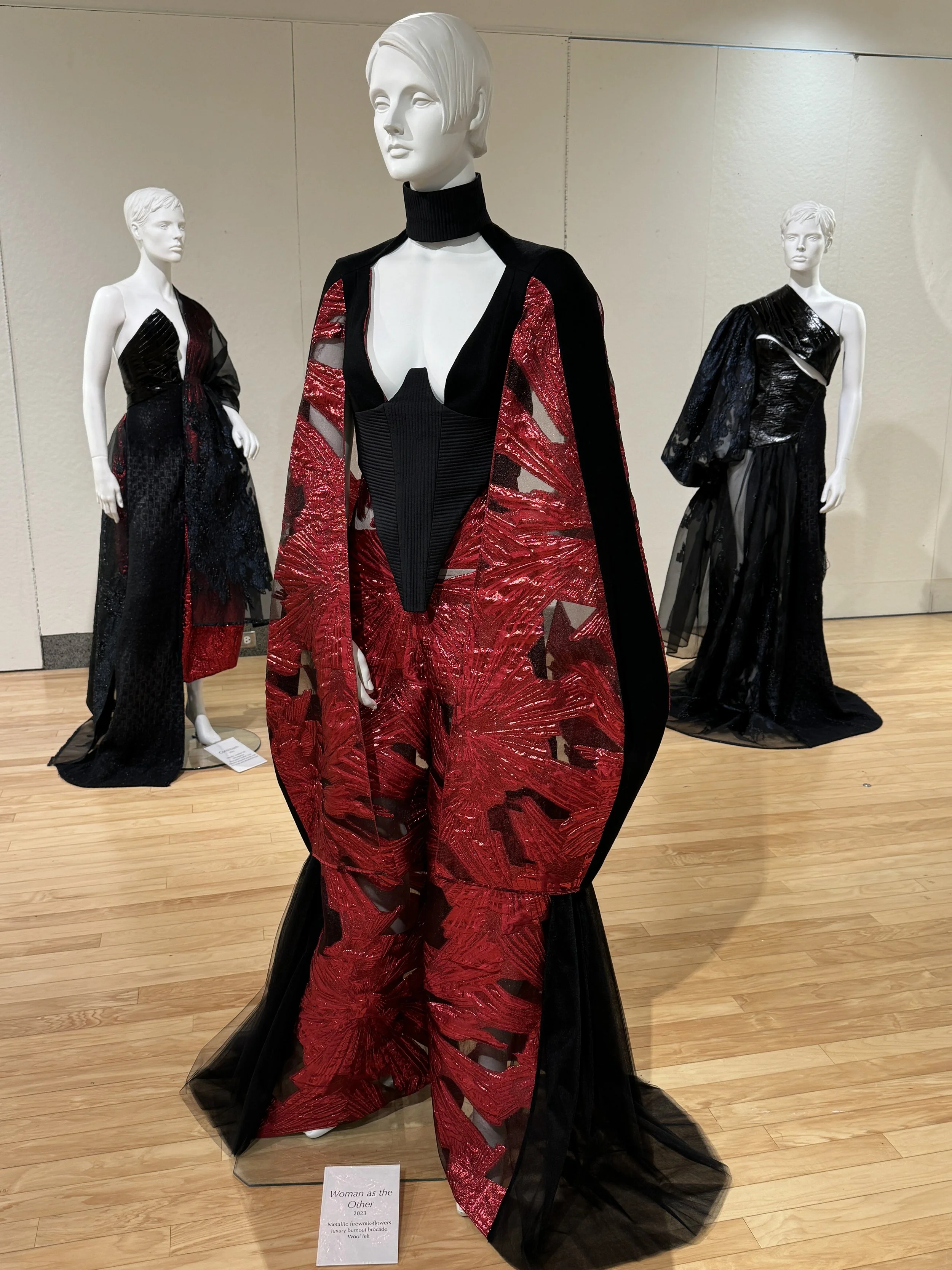 Three mannequins display avant-garde black and metallic red gowns in a fashion exhibition, including a dramatic red sequined dress with black tulle.