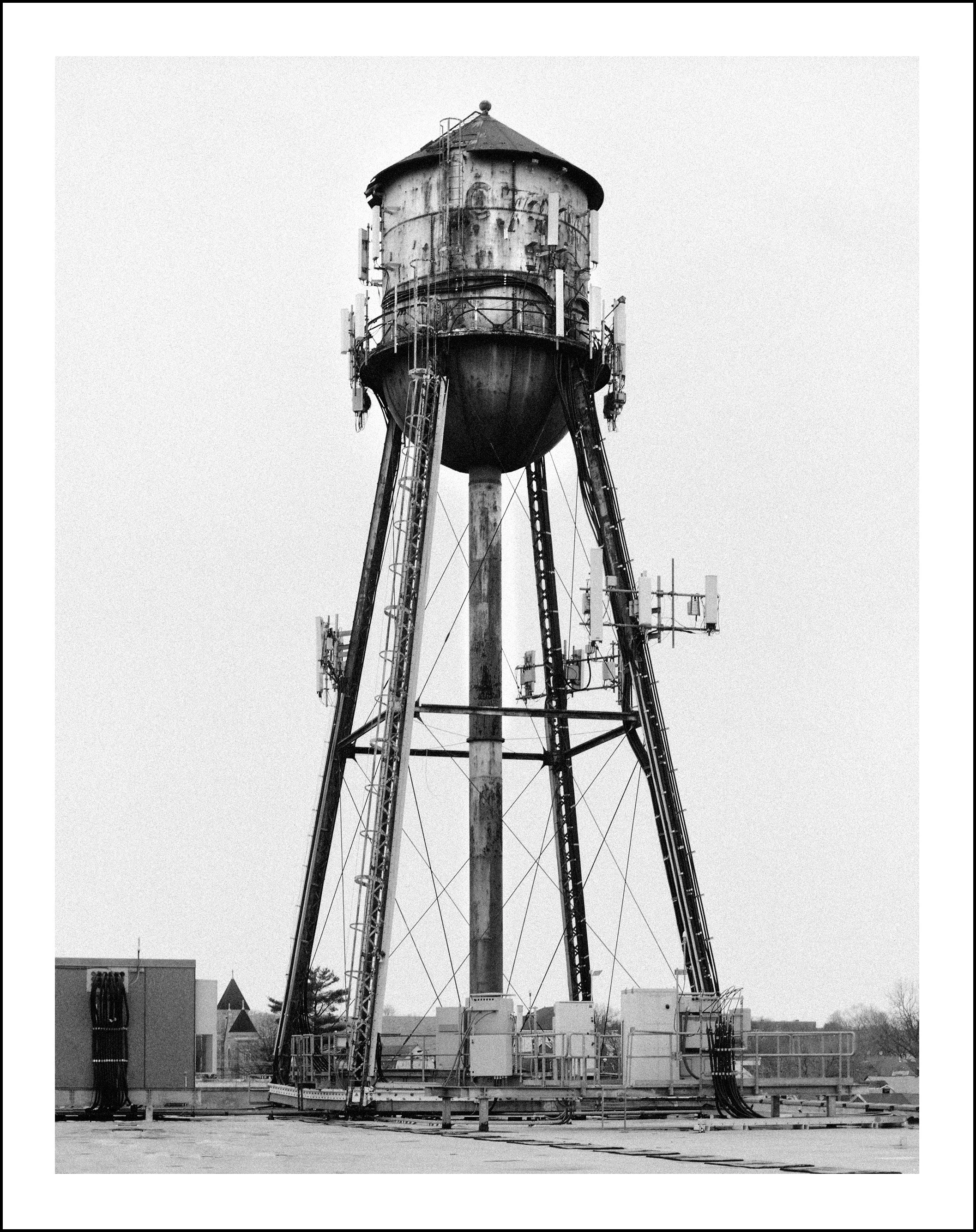 Black-and-white photo of a tall industrial water tower with metal support legs, ladders, cables, and antenna equipment.