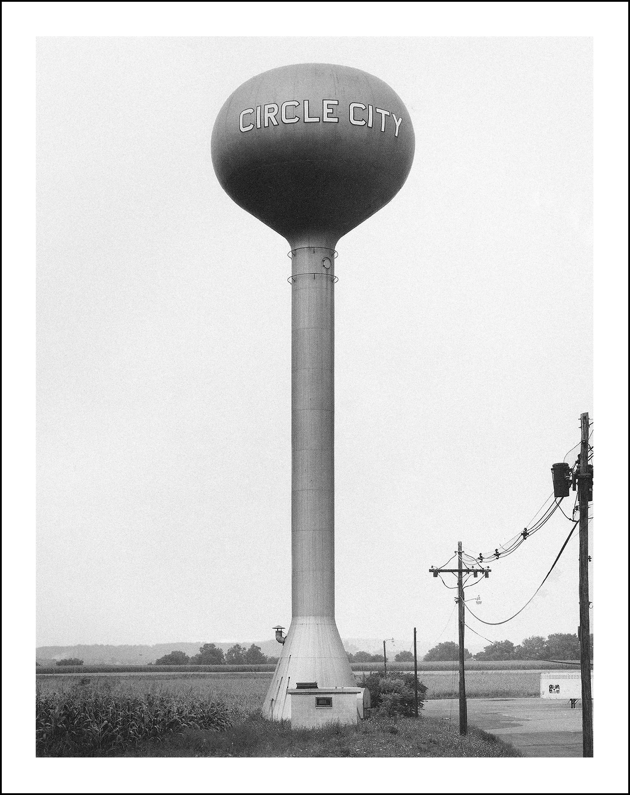 Black-and-white photo of the Circle City water tower beside a cornfield and utility poles