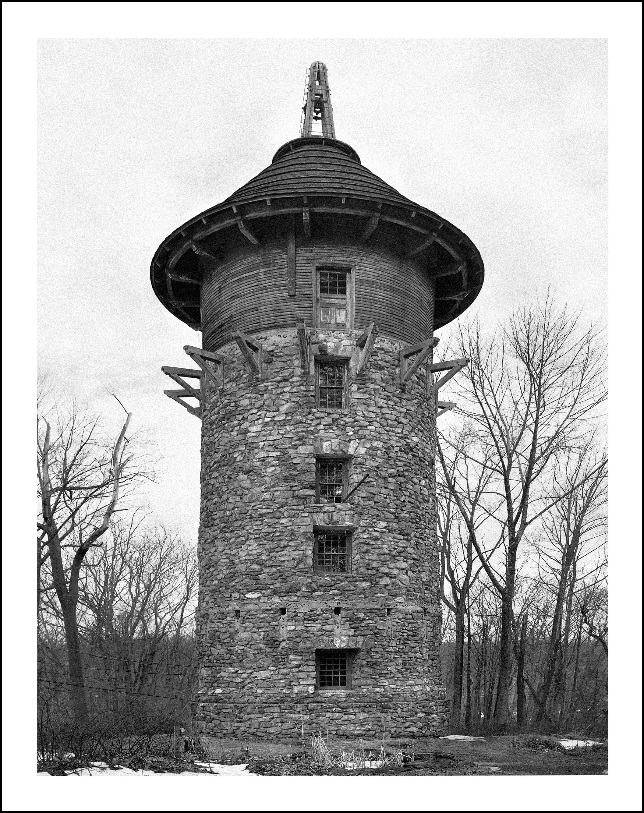 Black-and-white photo of a historic stone tower with a conical wooden roof and cupola in a leafless forest