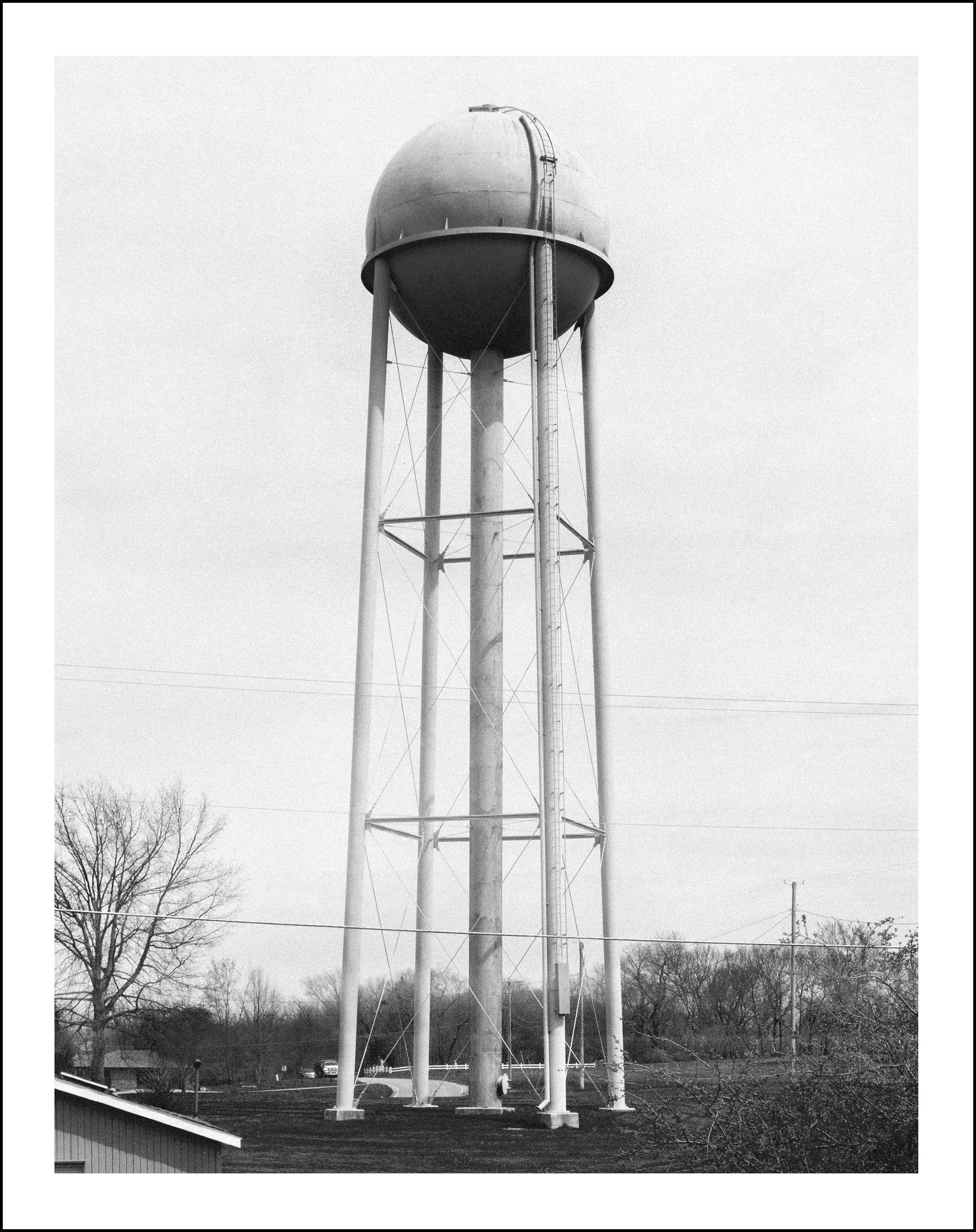 Black-and-white photograph of a tall elevated water tower with support legs, ladder, trees, and utility lines.