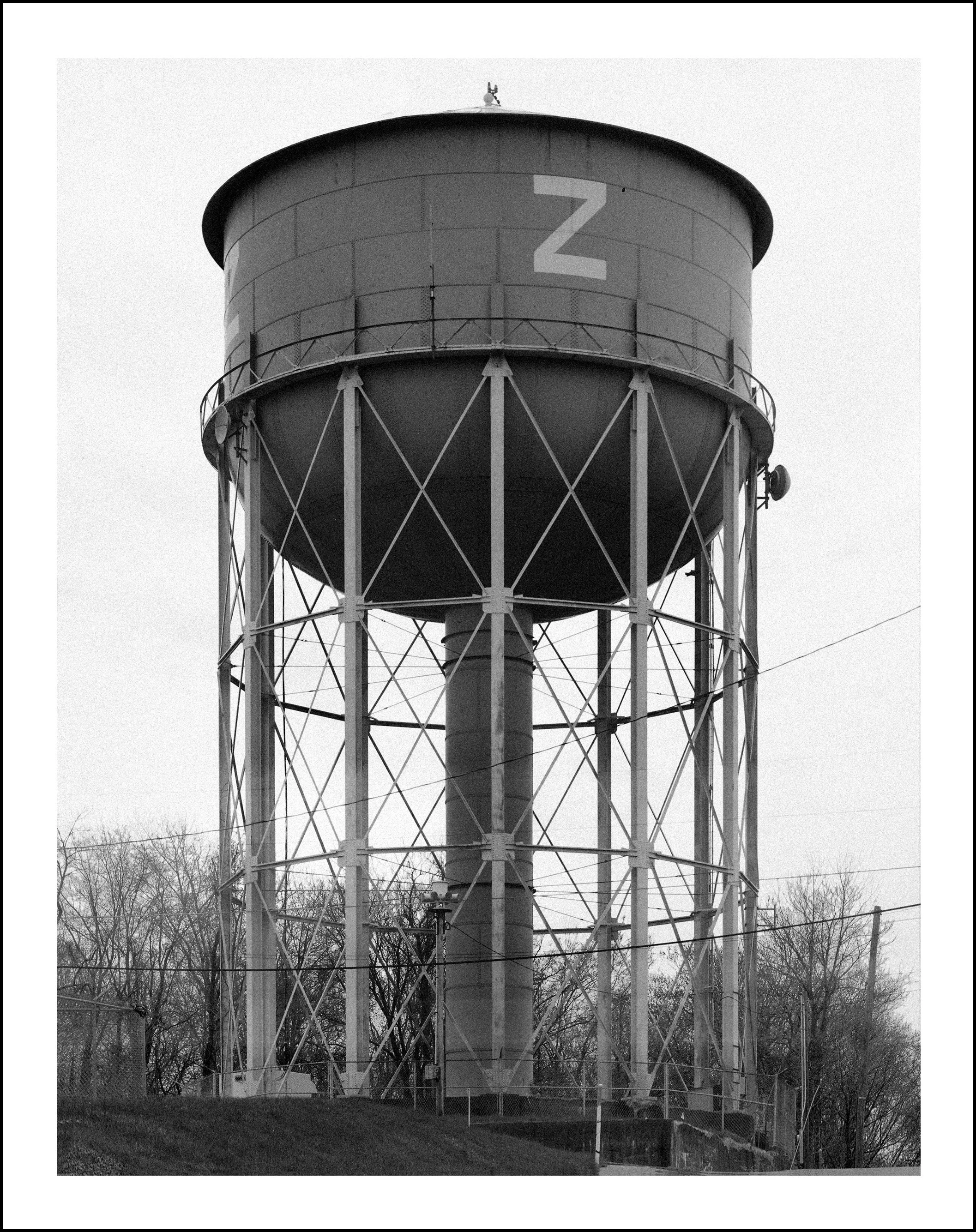 Black-and-white photo of a large elevated cylindrical water tower marked with the letter Z, surrounded by fencing and leafless trees.