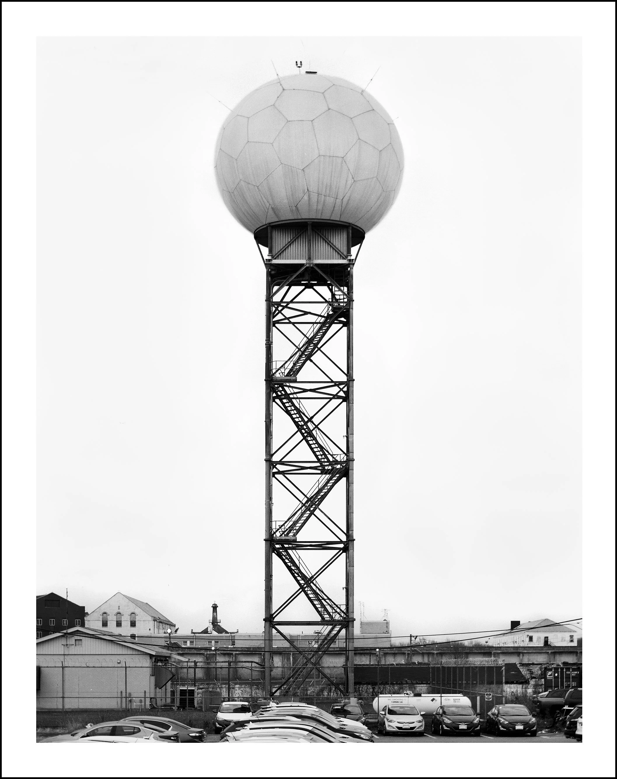 Black-and-white photo of a spherical water tower on a tall steel frame above an industrial parking lot.