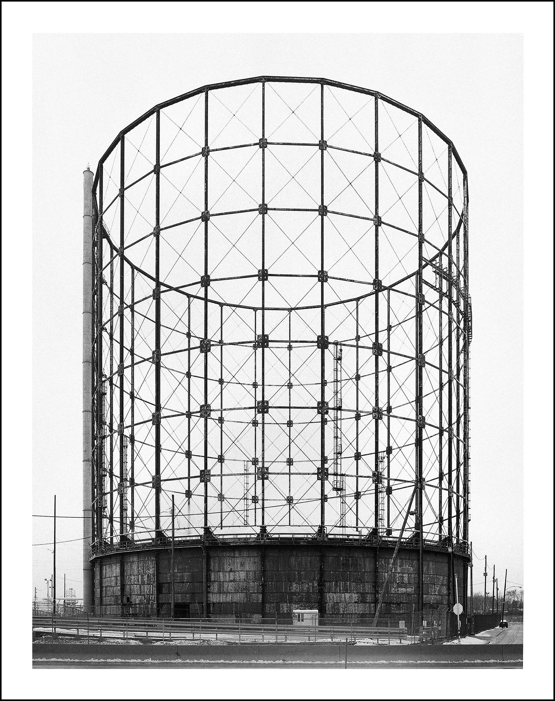Black-and-white photo of a large cylindrical steel gas holder with exposed lattice framework, industrial piping, and a smokestack.