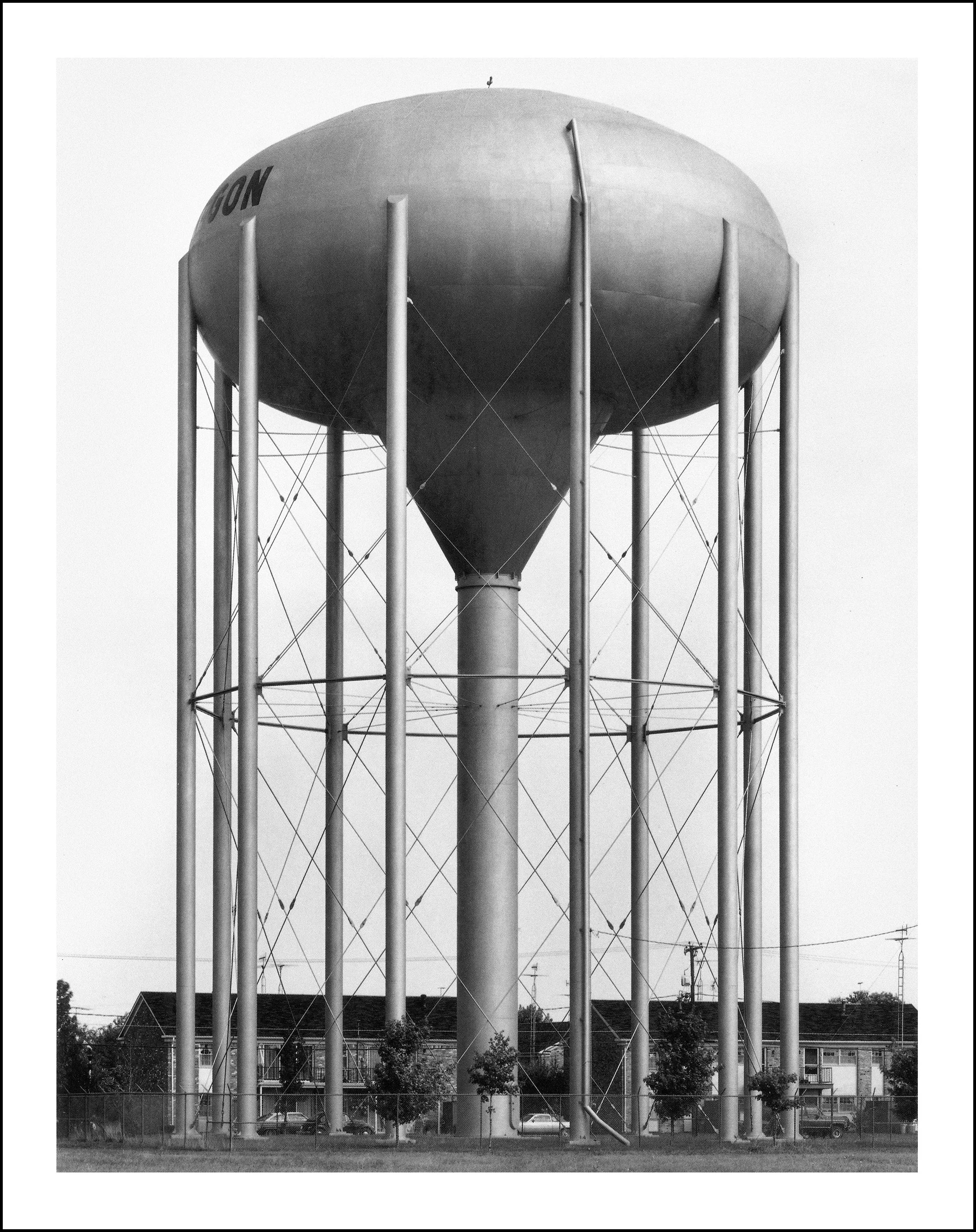 Black-and-white photograph of a large elevated water tower with cylindrical support legs, guy wires, and homes in the background.