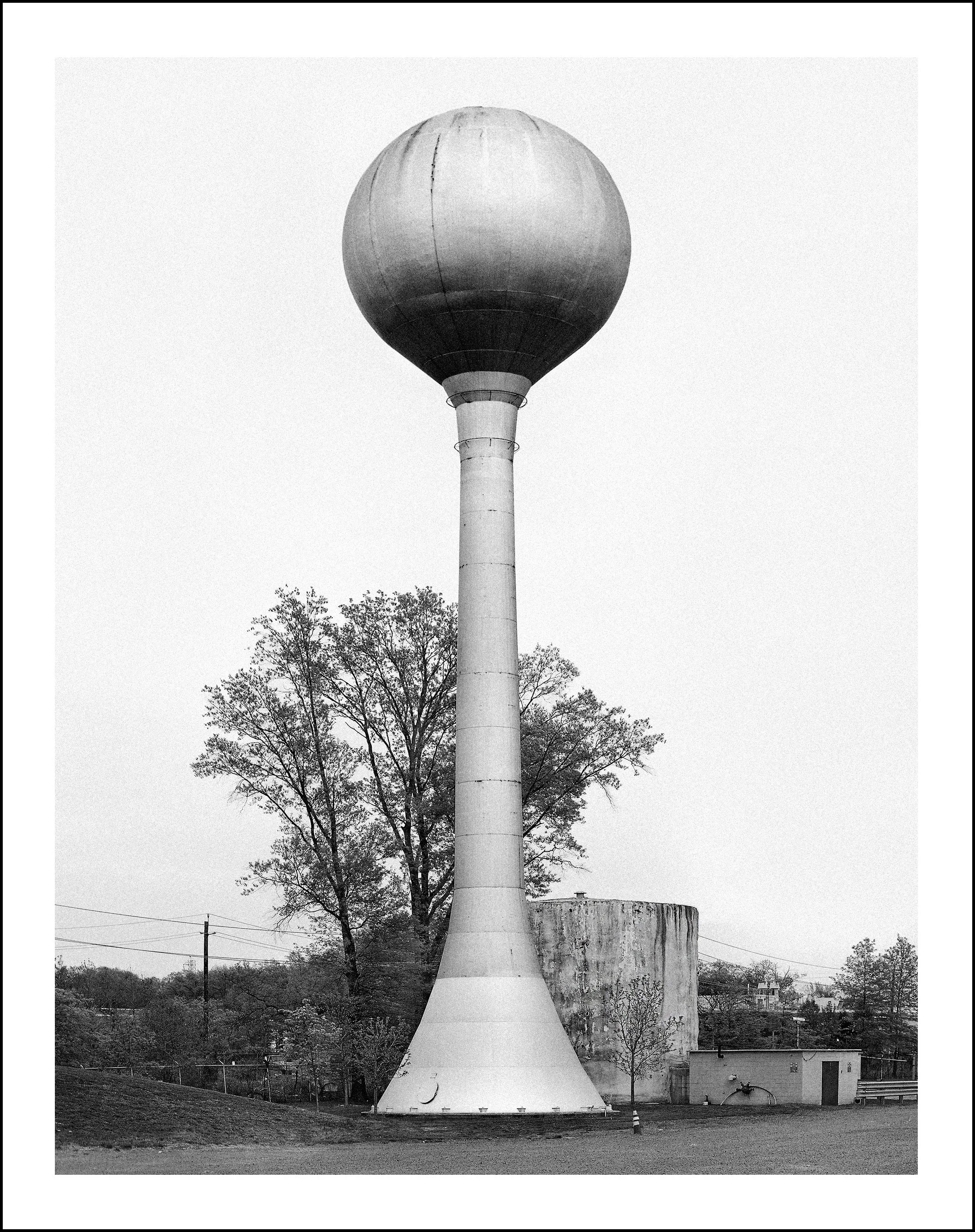 Black-and-white photograph of a large spherical water tower on a tall concrete column beside trees and utility buildings