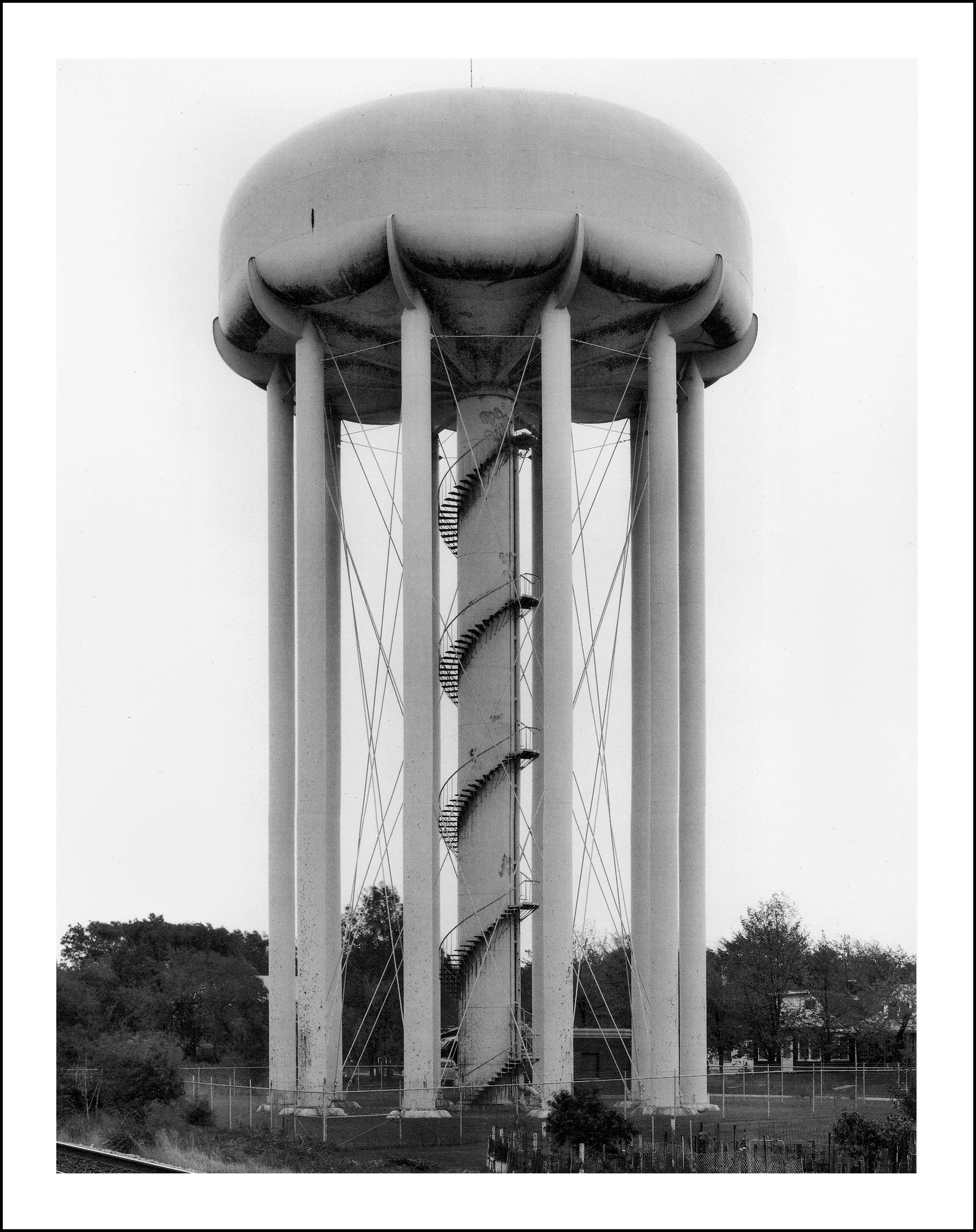 Black-and-white photograph of a tall water tower with a spiral staircase, support columns, and cables above a fenced industrial site.