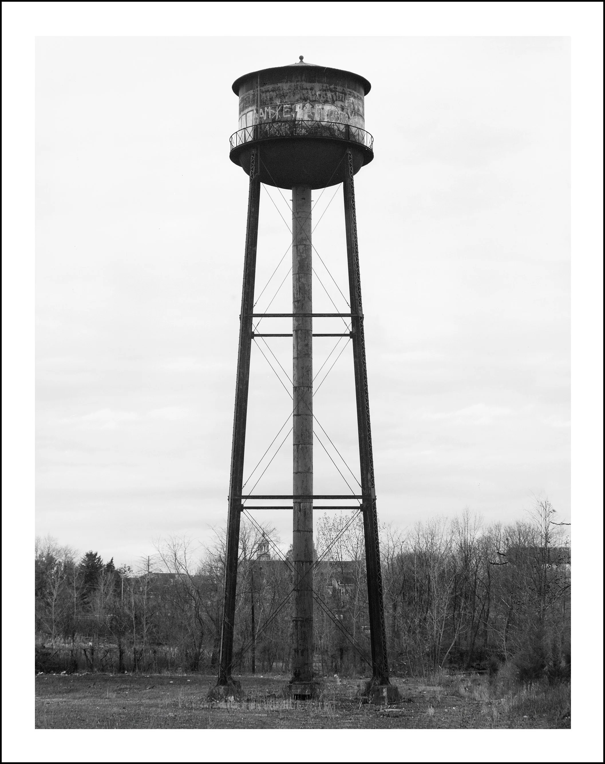 Black-and-white photograph of a tall, weathered water tower with graffiti, surrounded by leafless trees.