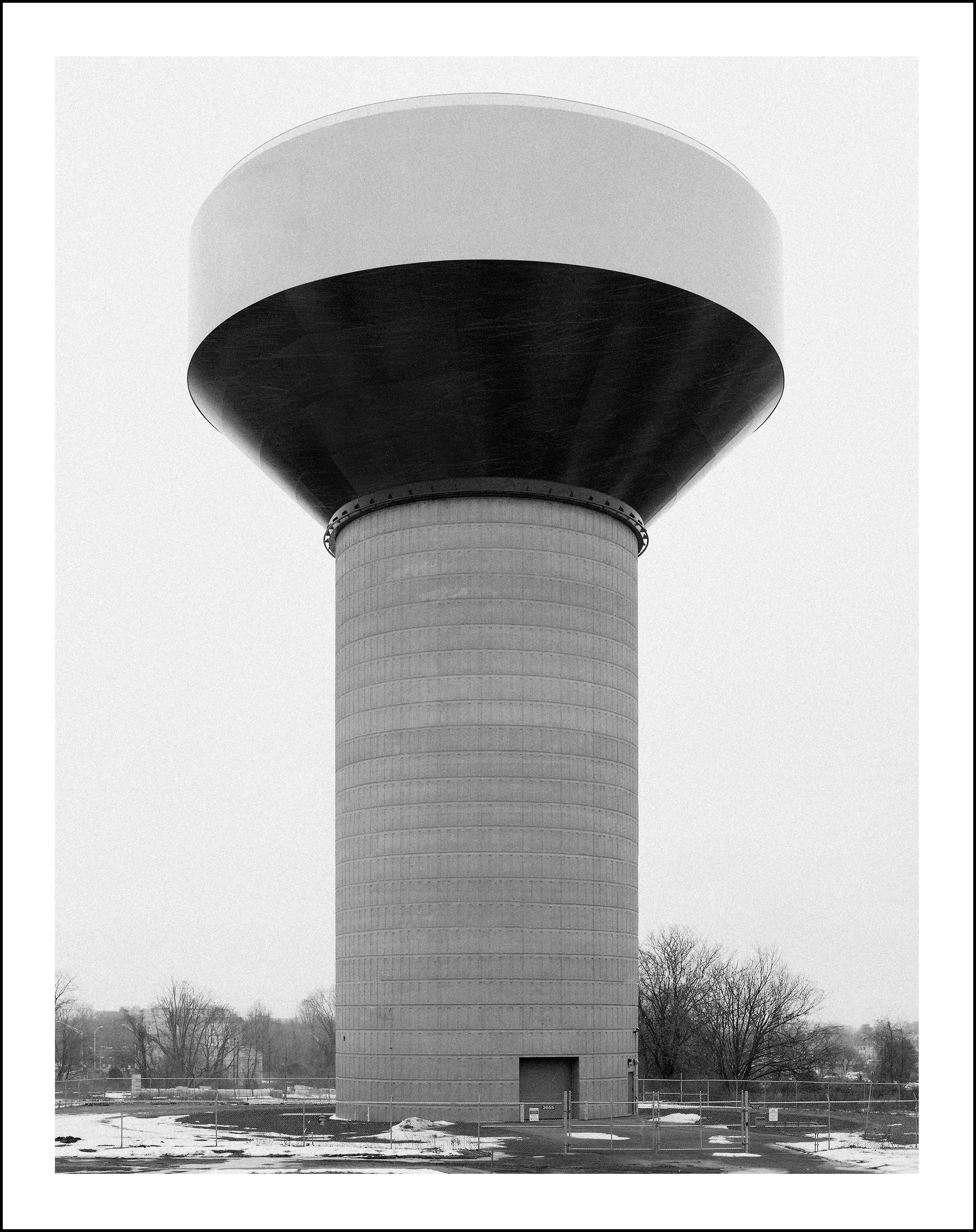 Black-and-white photo of a large concrete water tower with a mushroom-shaped roof in a snowy, fenced area