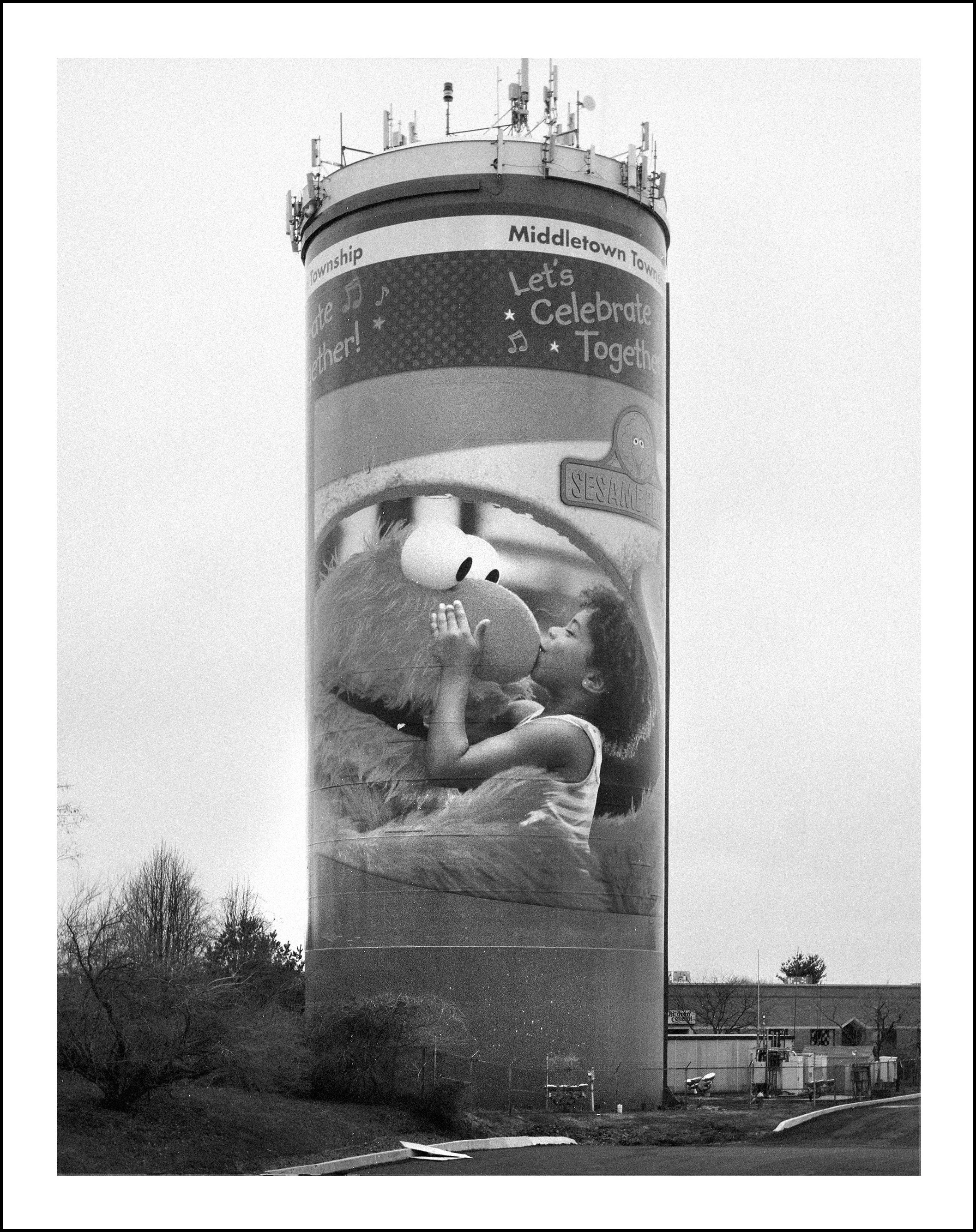 Black-and-white photo of a Middletown Township water tower decorated with a Sesame Street character hugging a child and the message “Let’s Celebrate Together.”