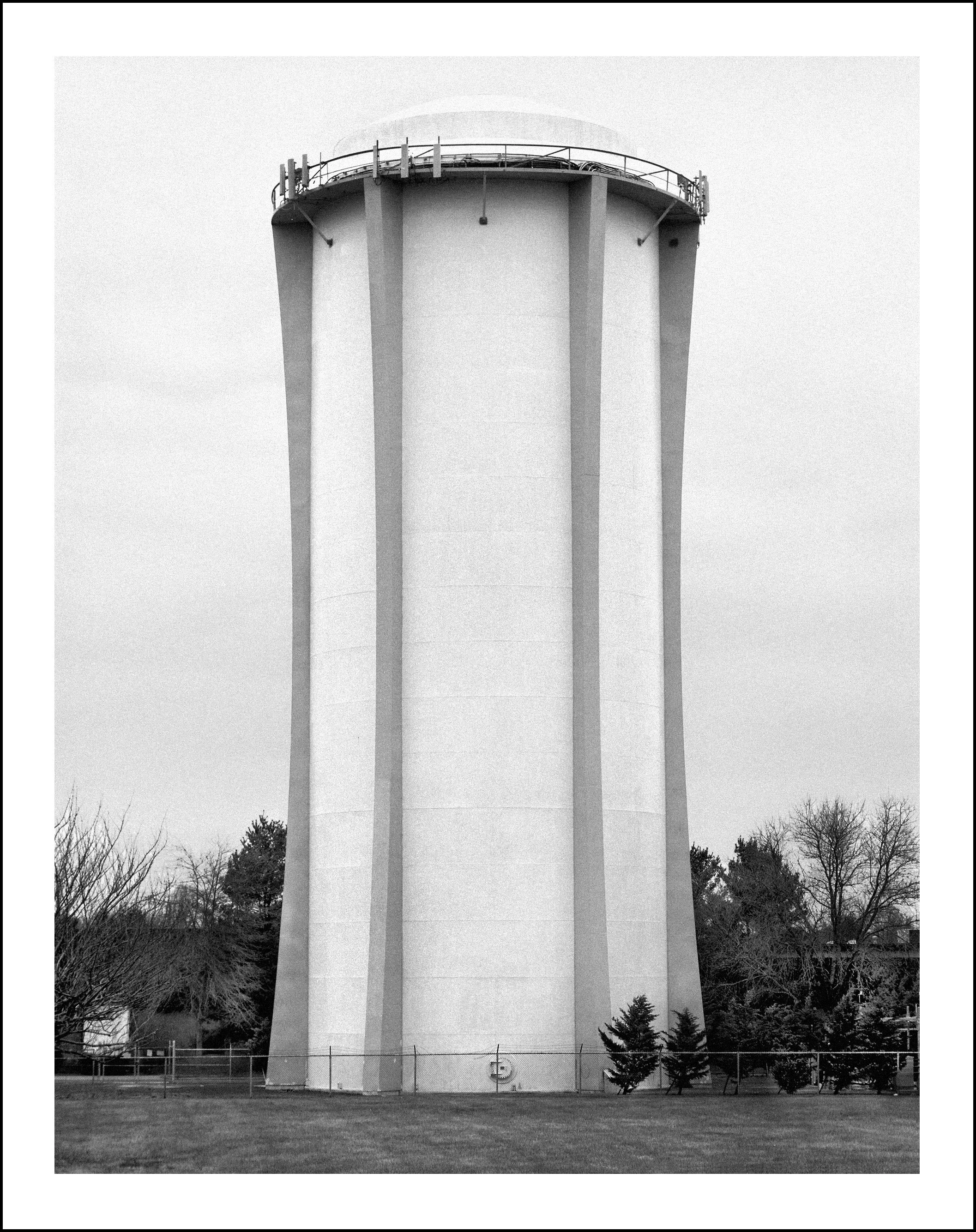 Black-and-white photo of a large cylindrical water tower with a ladder and rooftop railing, surrounded by trees and a chain-link fence