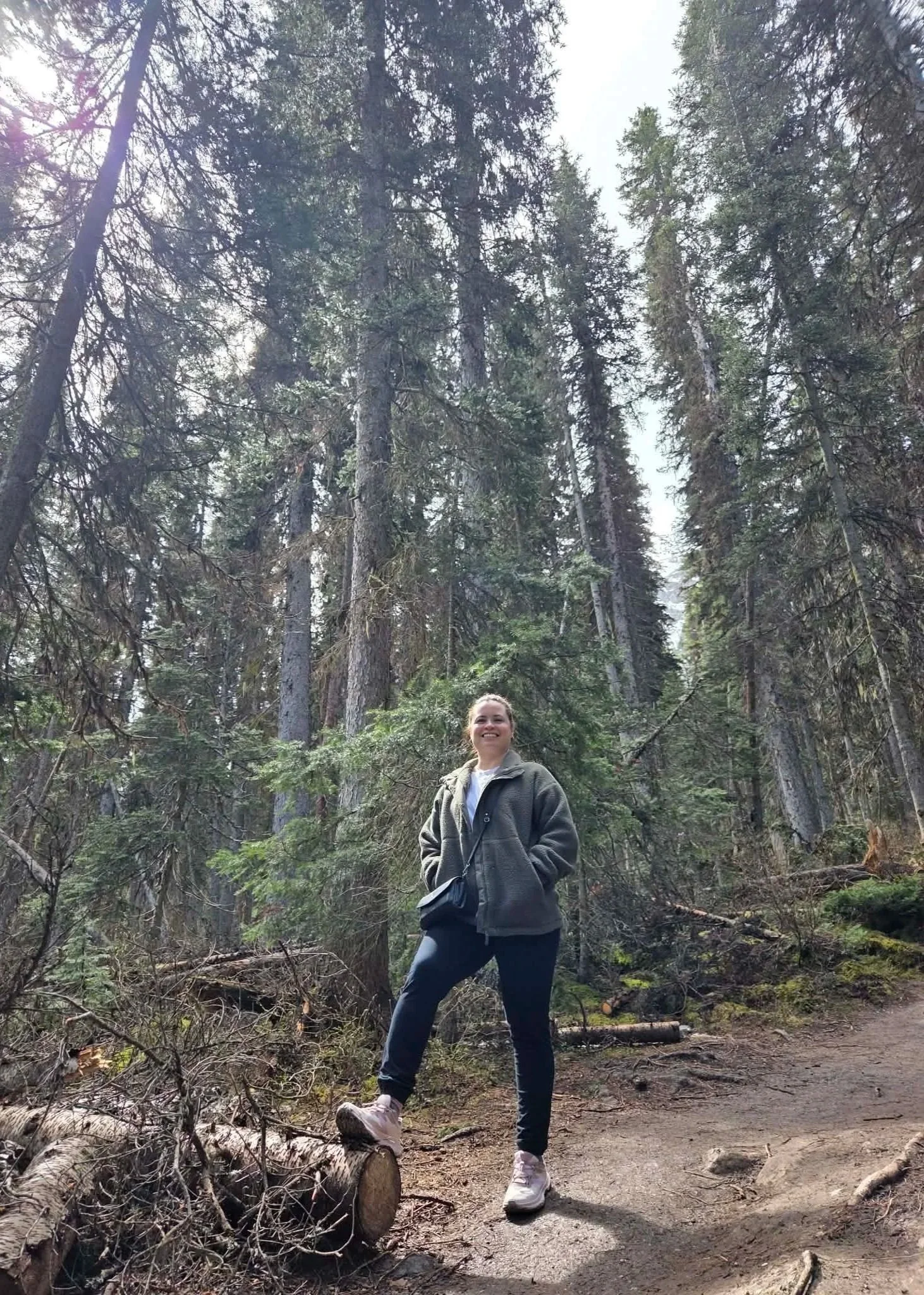 Smiling woman posing on a fallen log along a forest hiking trail surrounded by tall evergreen trees.