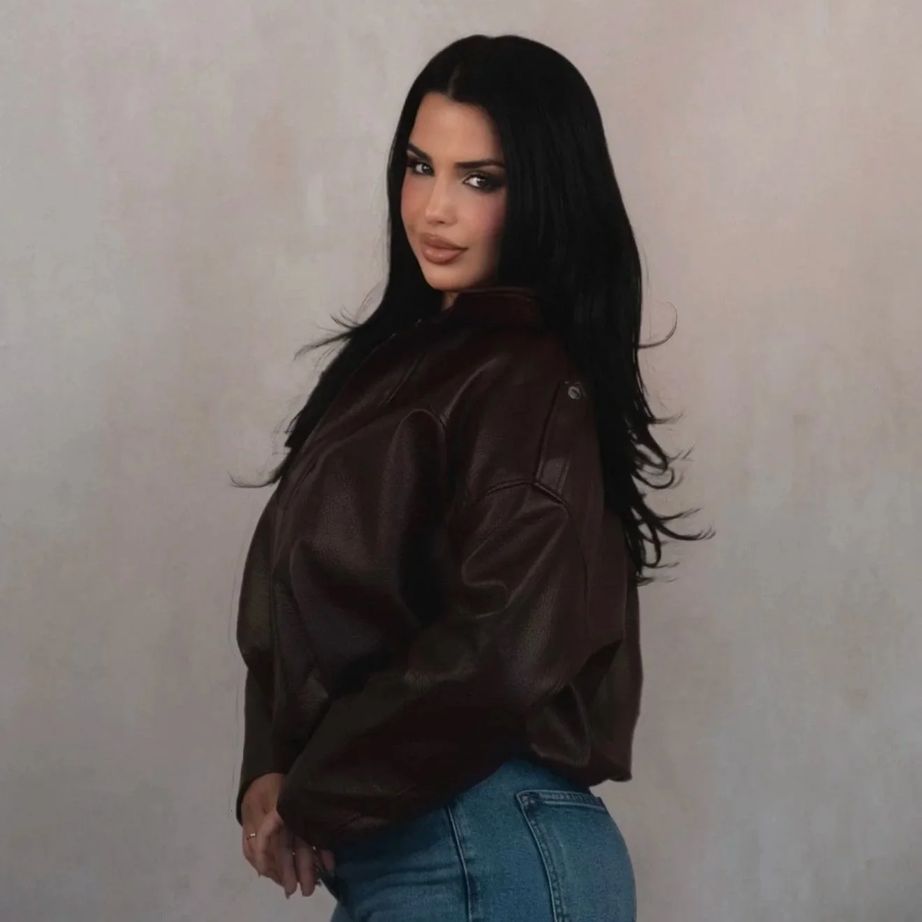 A woman with long black hair, wearing a dark leather jacket and blue jeans, posing against a plain light-colored wall.