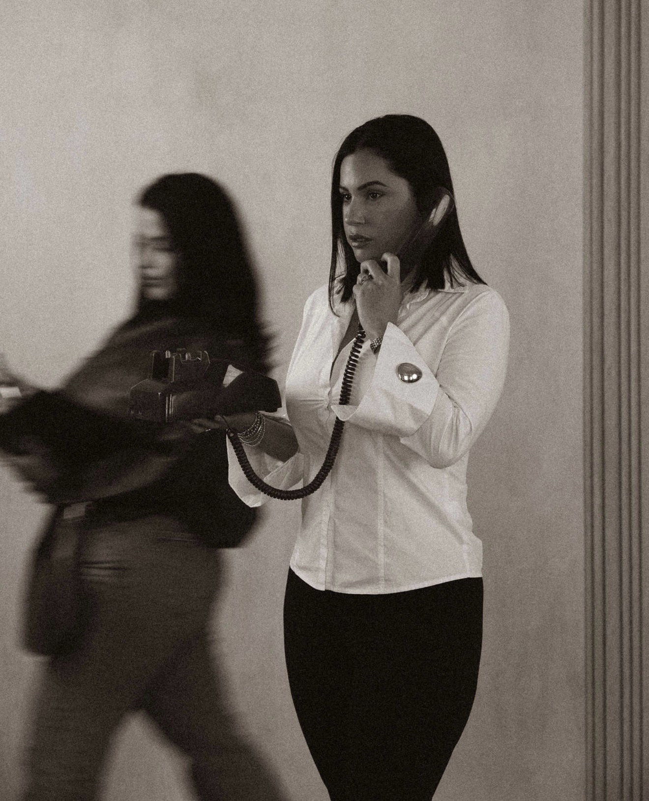 A woman in a white shirt holding a vintage telephone receiver, looking focused. A second blurred woman is walking past in the foreground.