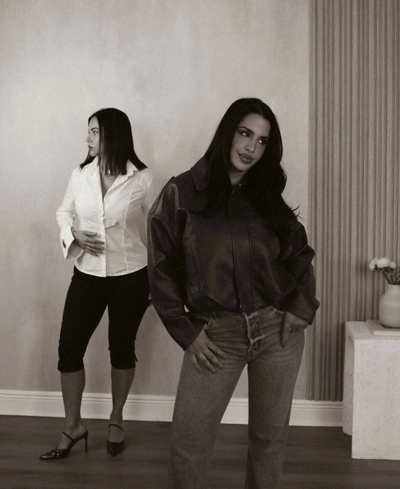 Two women with dark hair and light skin posing indoors. The woman in the foreground is wearing a dark leather jacket and jeans, while the woman in the background is in a white blouse and black pants. The background features a plain wall and a side table with a vase of flowers.