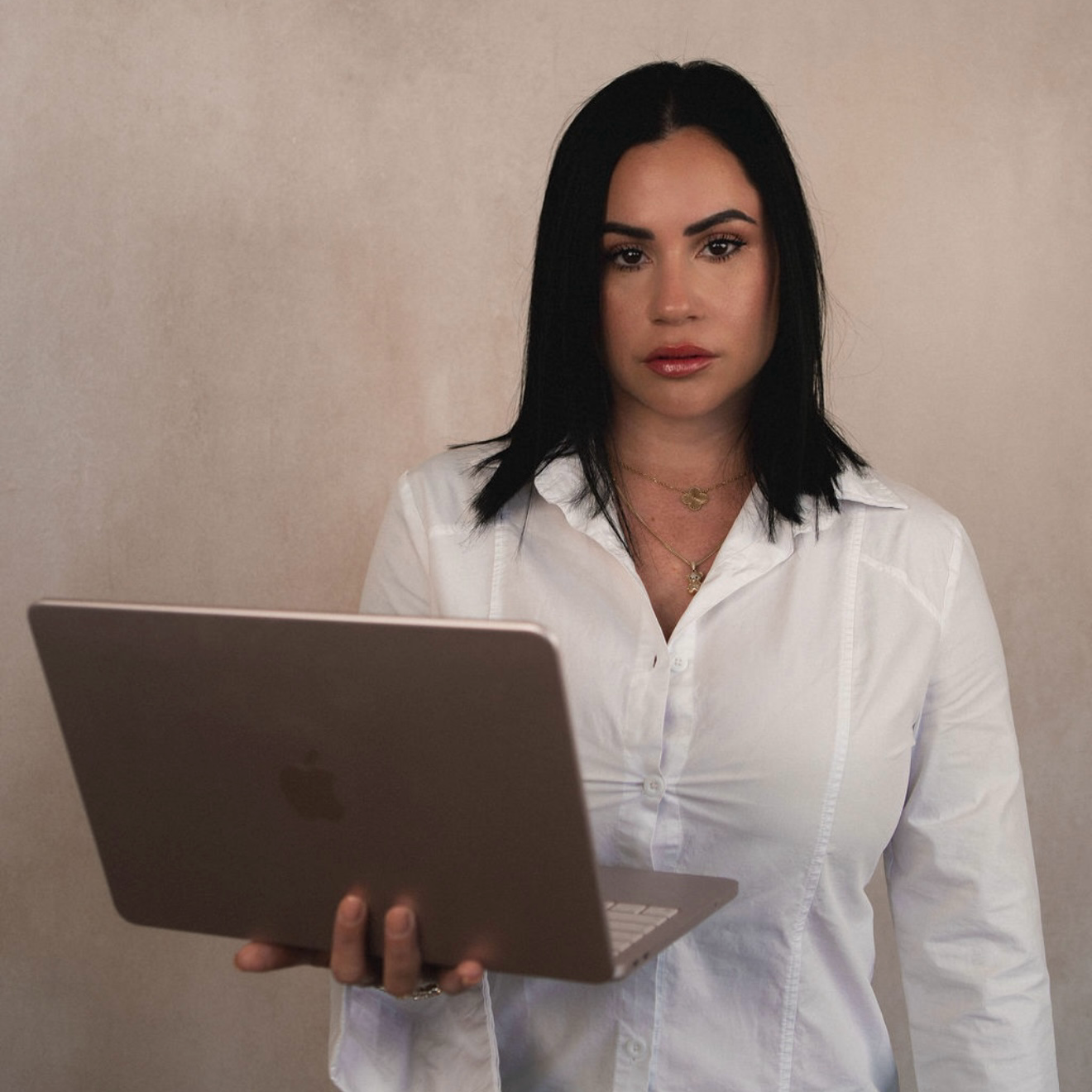 A woman with black hair wearing a white shirt, holding a silver laptop, standing against a plain beige wall.