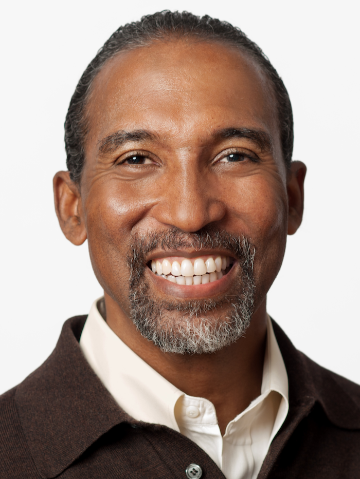 Close-up portrait of a middle-aged man with a big smile, gray beard, and mustache, wearing a white shirt and dark sweater, against a plain white background.