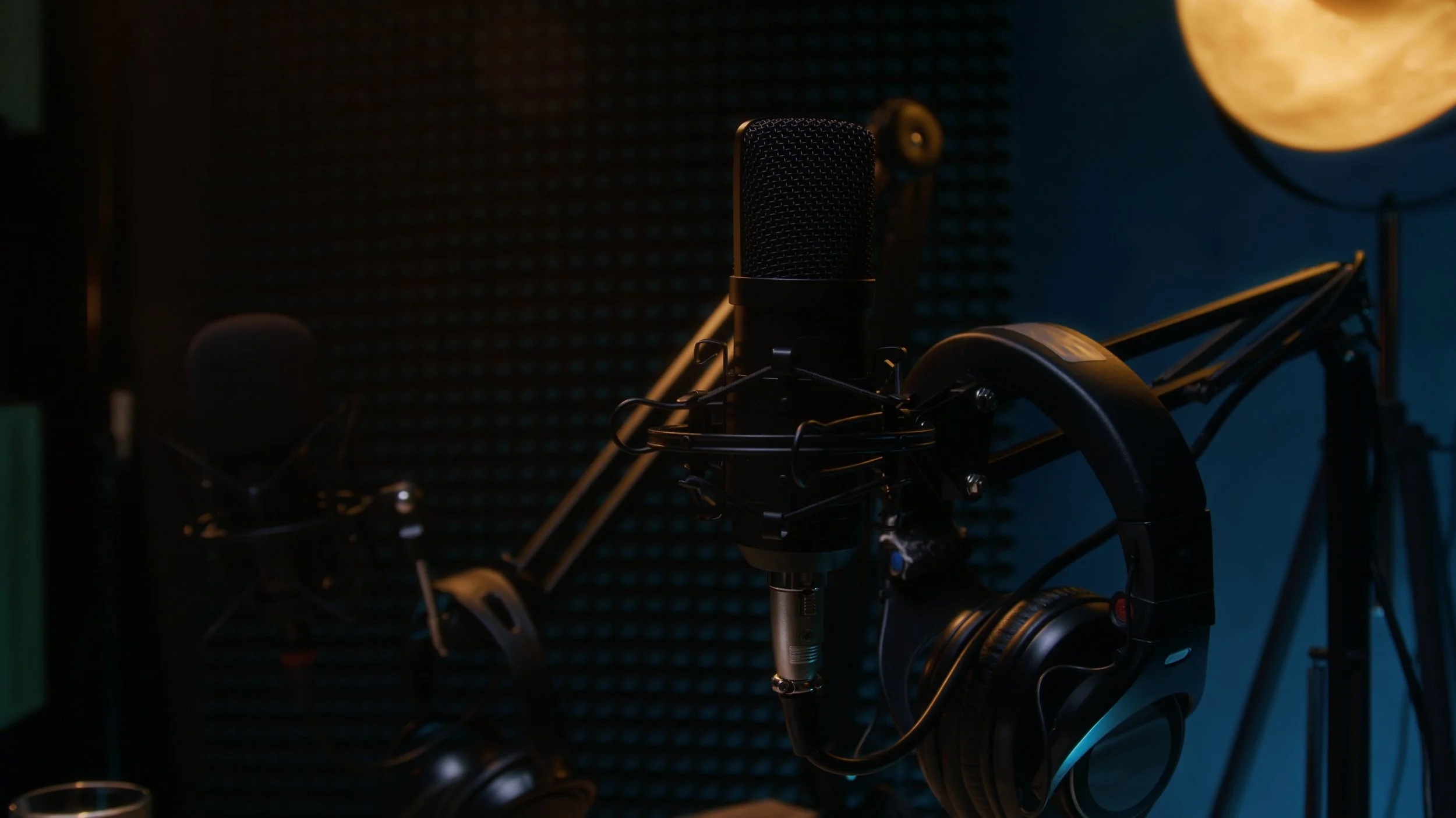 A recording studio with a professional microphone mounted on a stand, surrounded by headphones, in a dimly lit room with soundproofing foam walls and a large moon-shaped lamp in the background.
