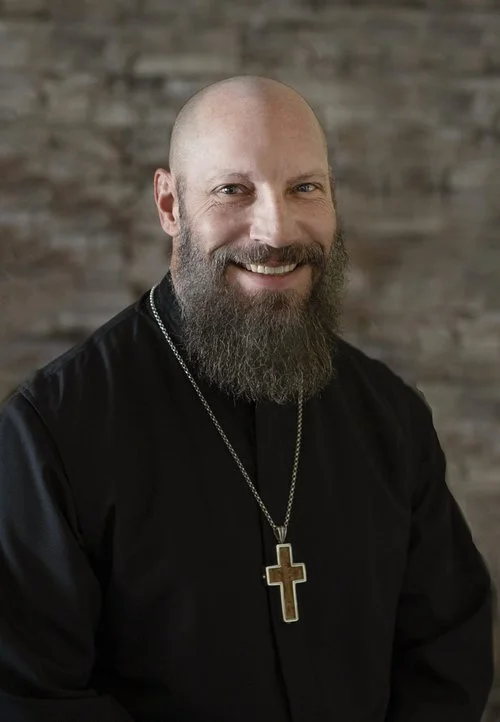 Fr. Timothy smiling, wearing a black clerical shirt and a wooden pectoral cross, in front of a brick wall