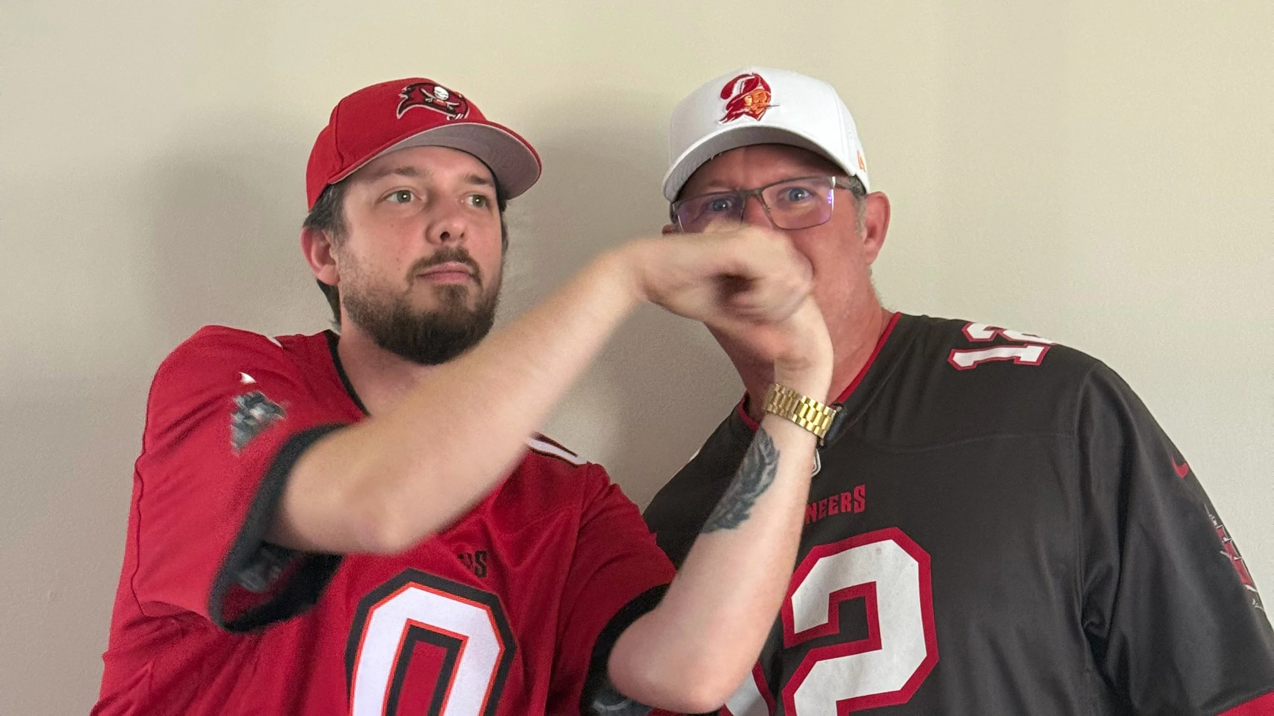 Two men wearing sports jerseys and baseball caps with Tampa Bay Buccaneers logos. One man has a beard and short dark hair, and the other is wearing glasses, has a tattoo on his arm, and is wearing a gold watch. They are making a fist bump.