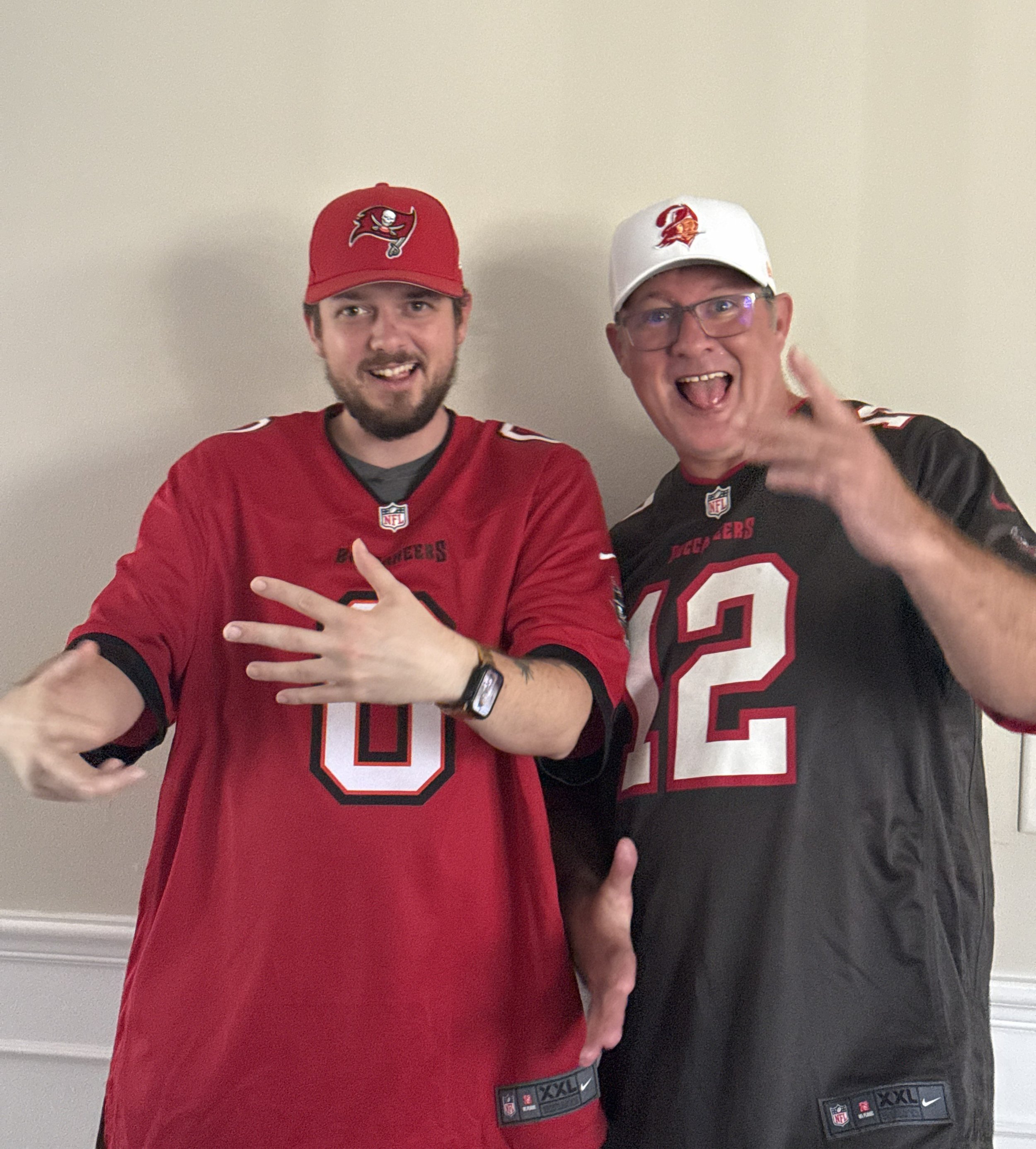 Two men wearing NFL football jerseys and hats, smiling and making gestures, standing against a plain wall.