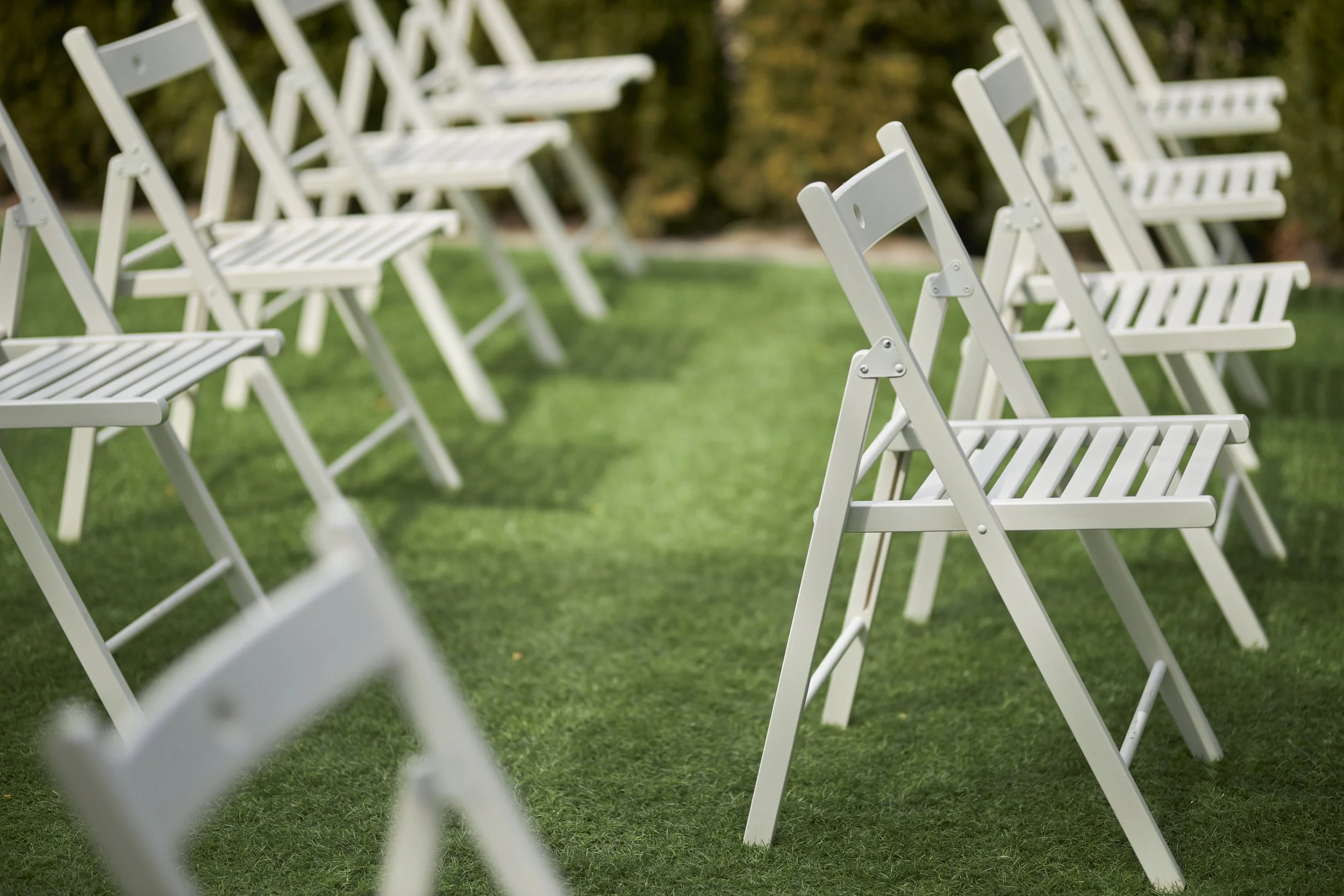 Multiple white folding chairs arranged outdoors on a grassy area, with trees in the background.