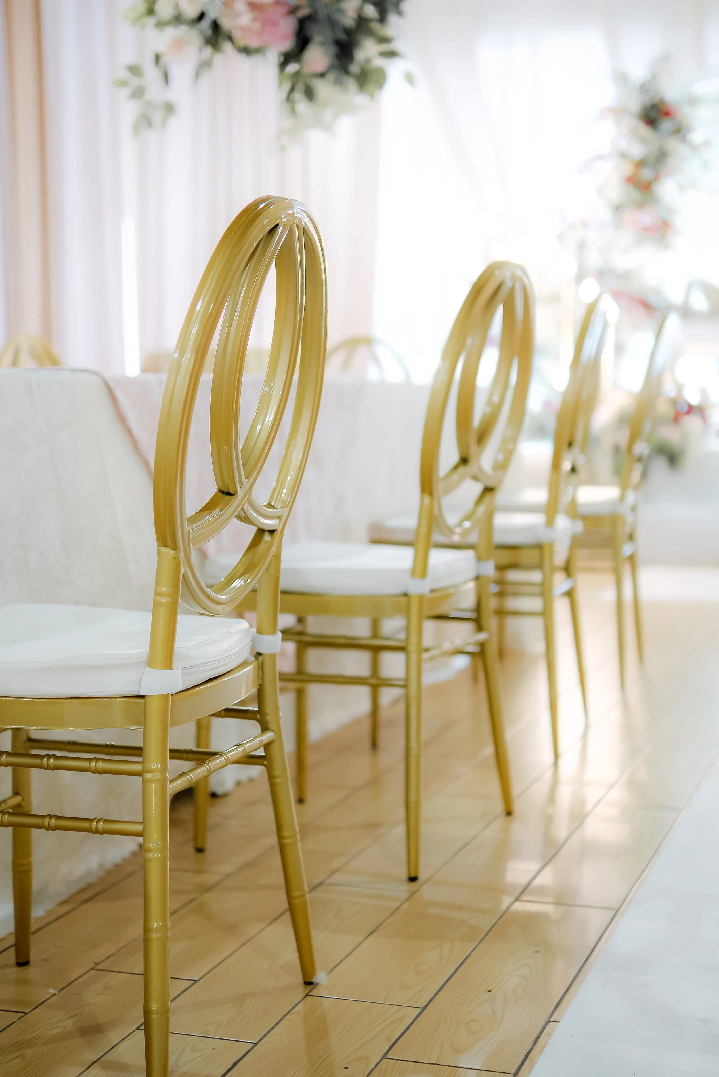Row of gold-colored chairs with white cushions around a decorated dining table, with floral arrangements and curtains in the background.